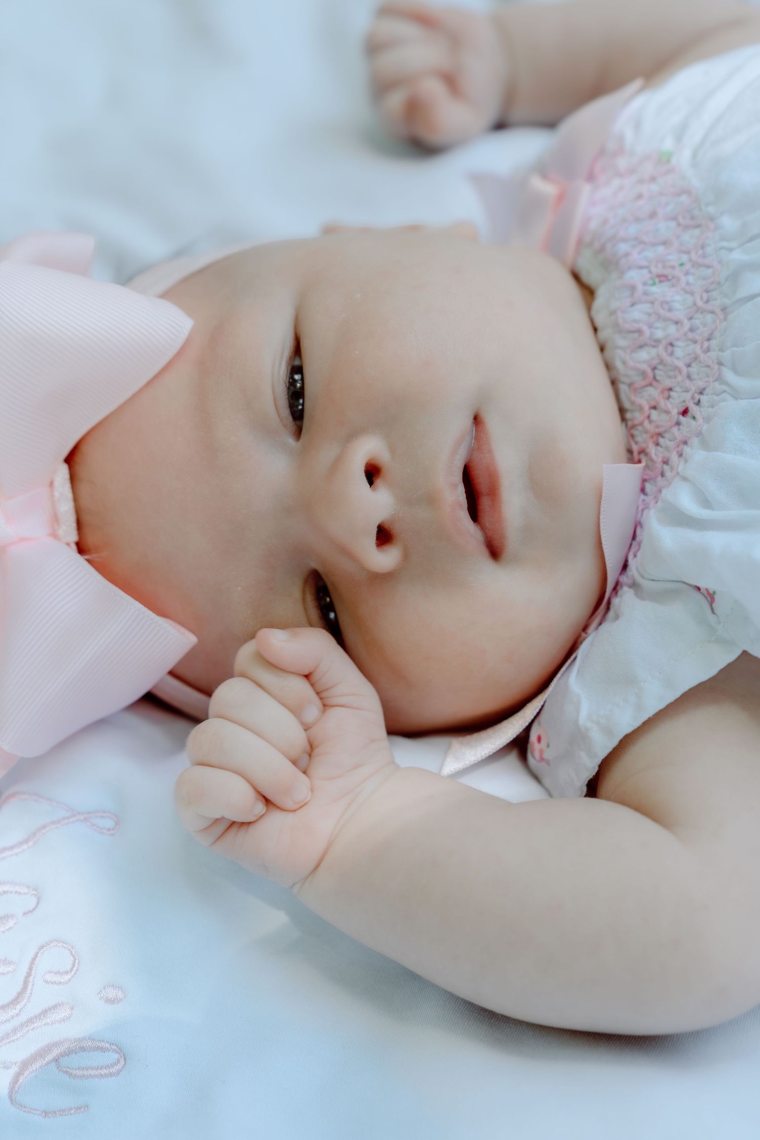 Alert newborn baby girl with dark eyes wearing pink bow headband lying on white embroidered blanket.