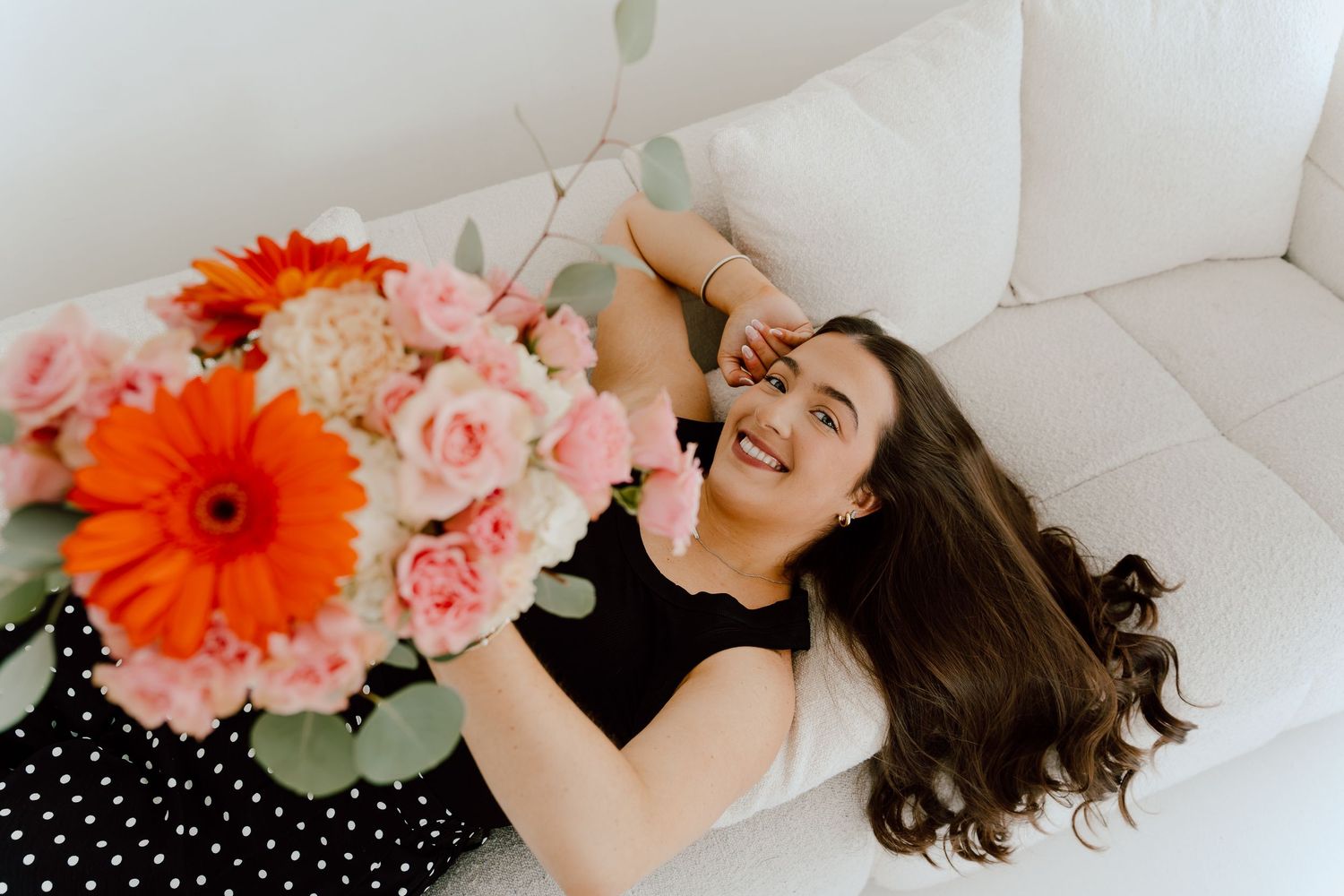 Laughing woman lying on white sofa with a vibrant bouquet of orange and pink flowers overhead.