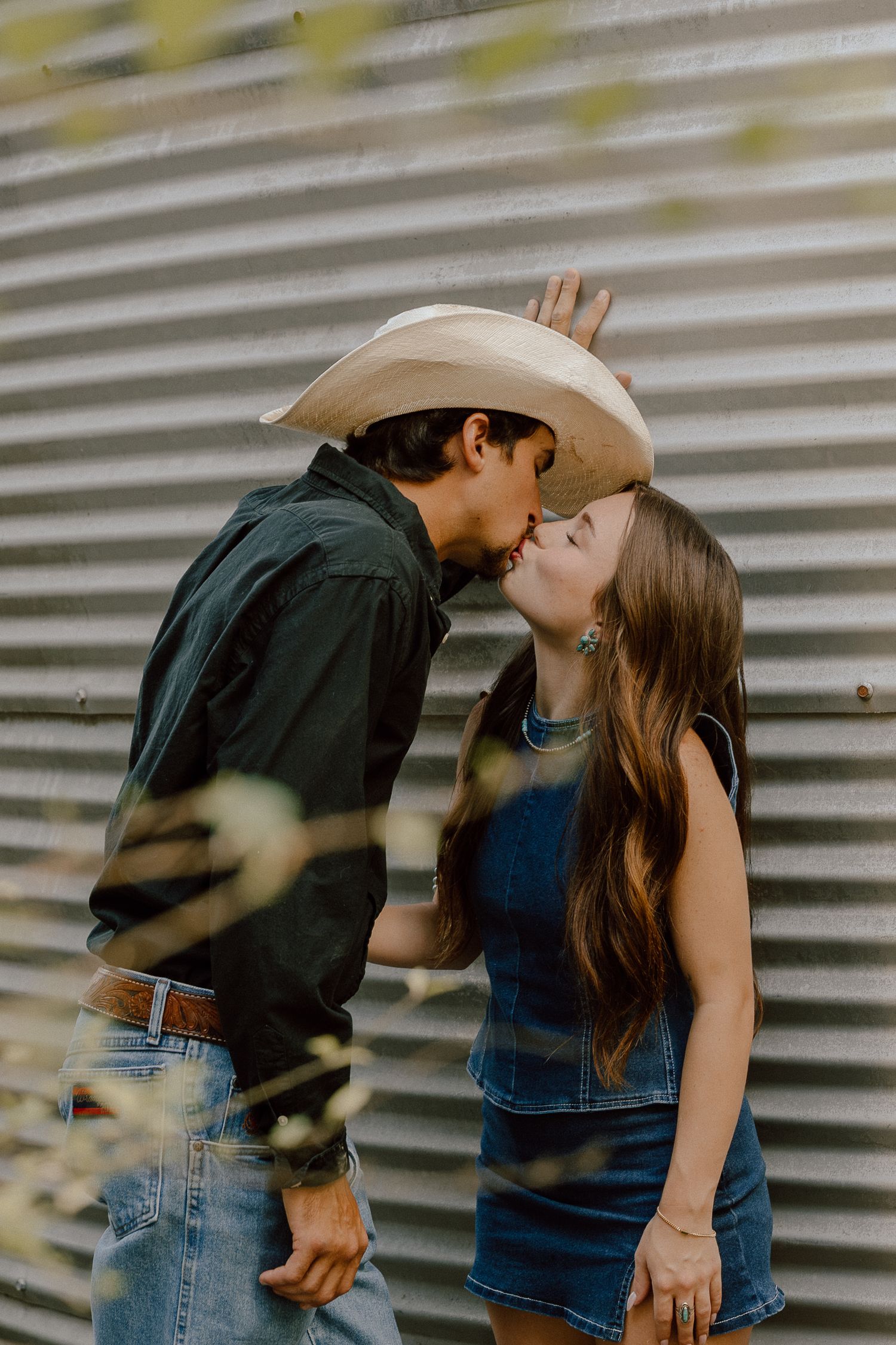 Couple sharing a kiss outdoors, man wearing cowboy hat and black shirt, woman in denim dress against corrugated metal wall.