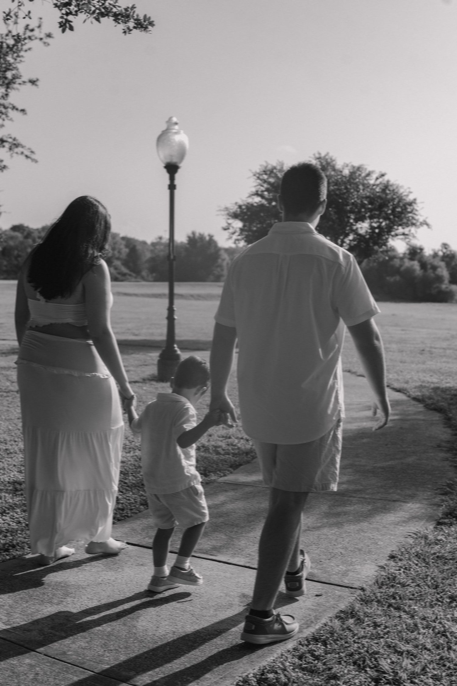 Black and white photo of a family walking hand in hand with their toddler on a park path.