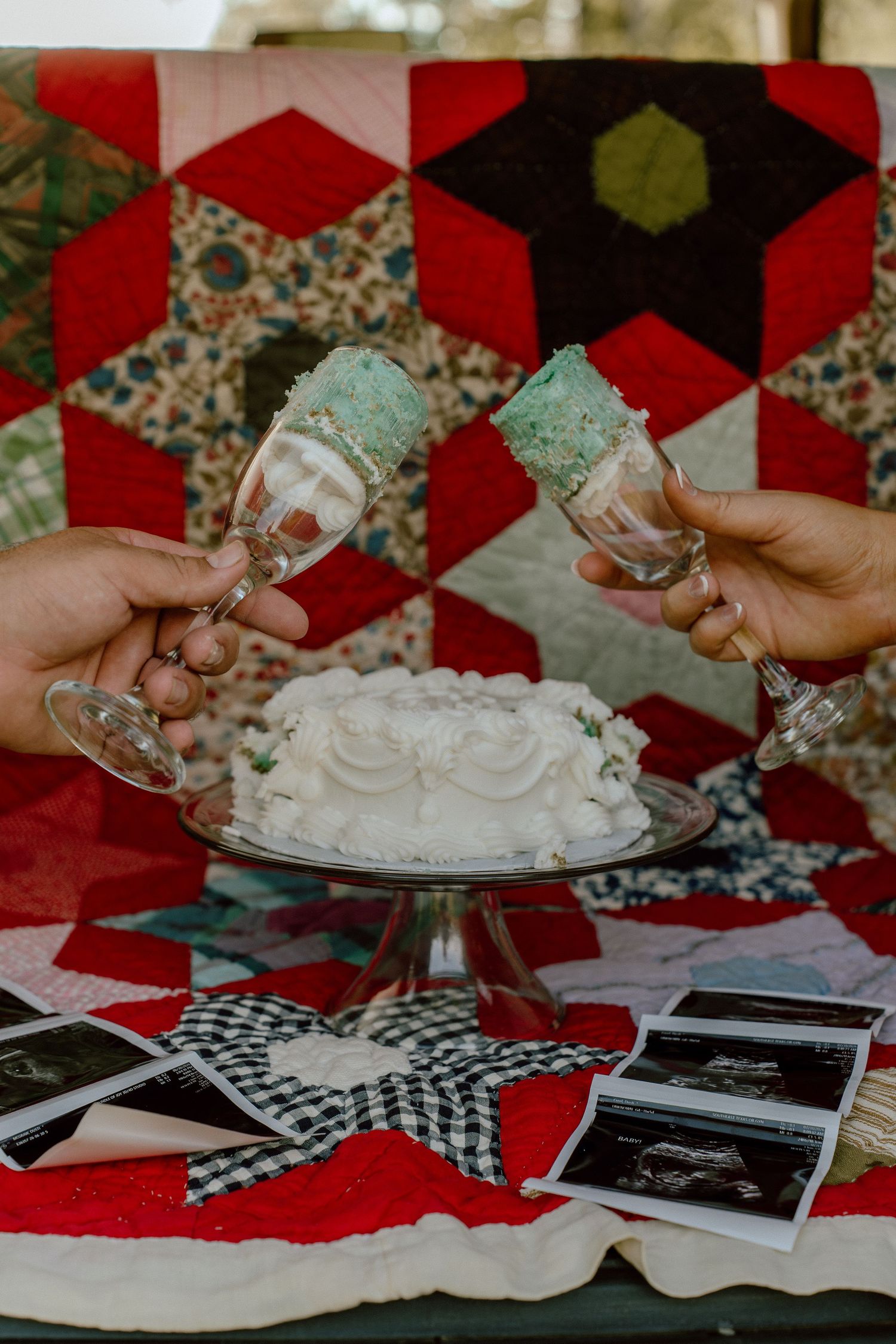 Two hands toasting with mint-rimmed champagne flutes over a white cake, with ultrasound photos and a colorful quilt backdrop.