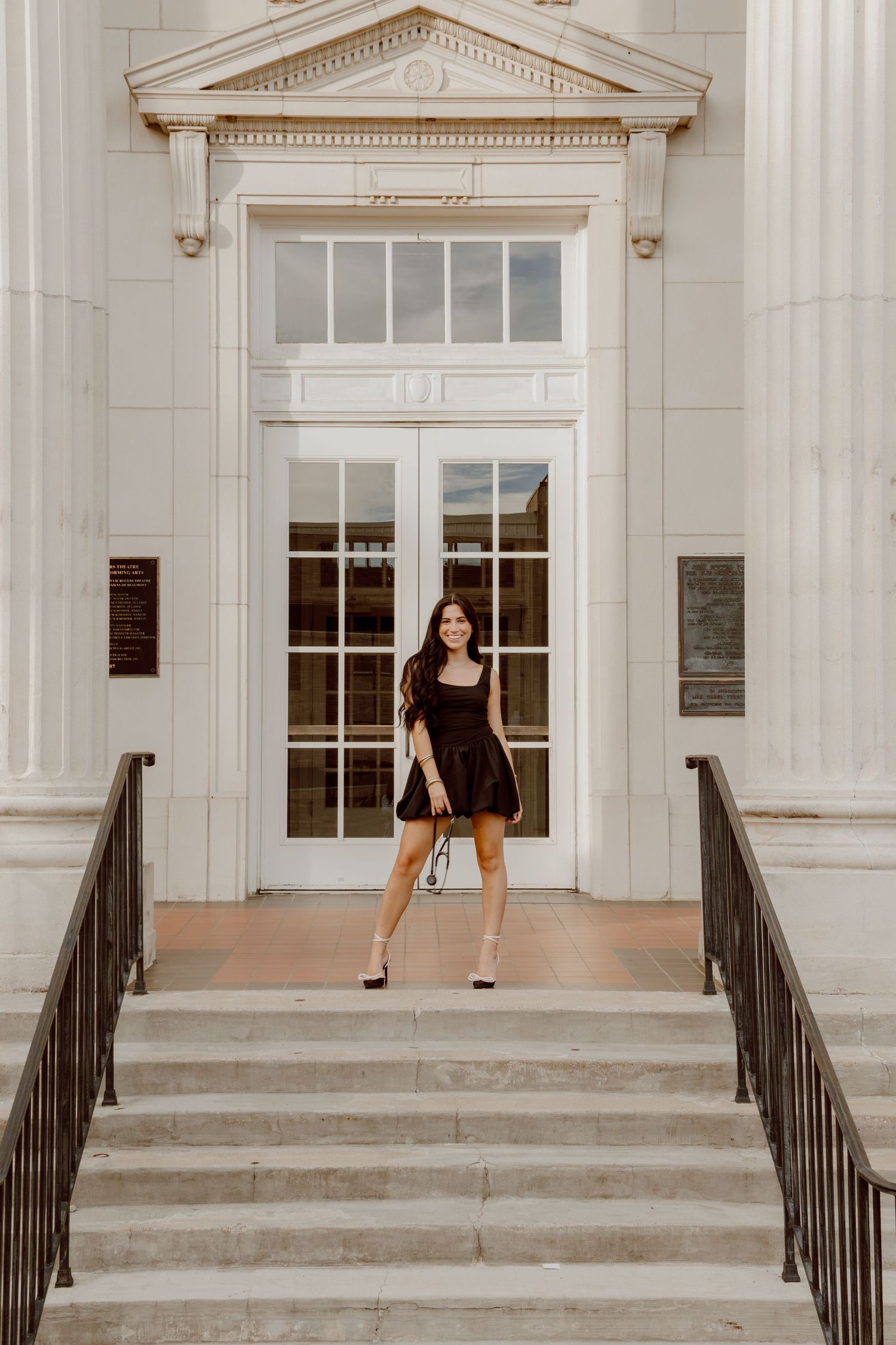 Brunette woman in black mini dress posing on steps of elegant white building for senior photos.
