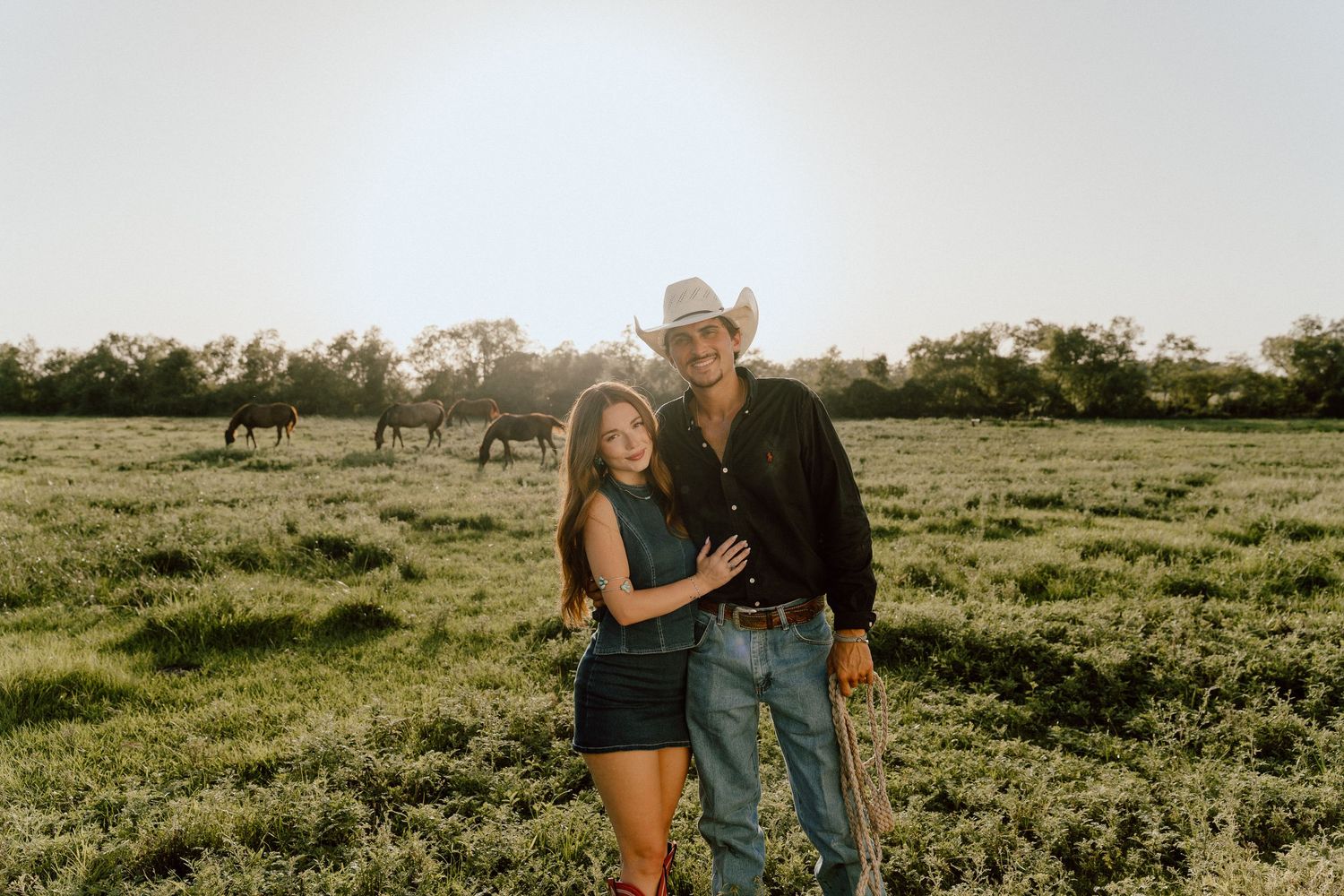 Couple embracing in a sunlit green pasture with horses grazing in the background, man wearing a cowboy hat.