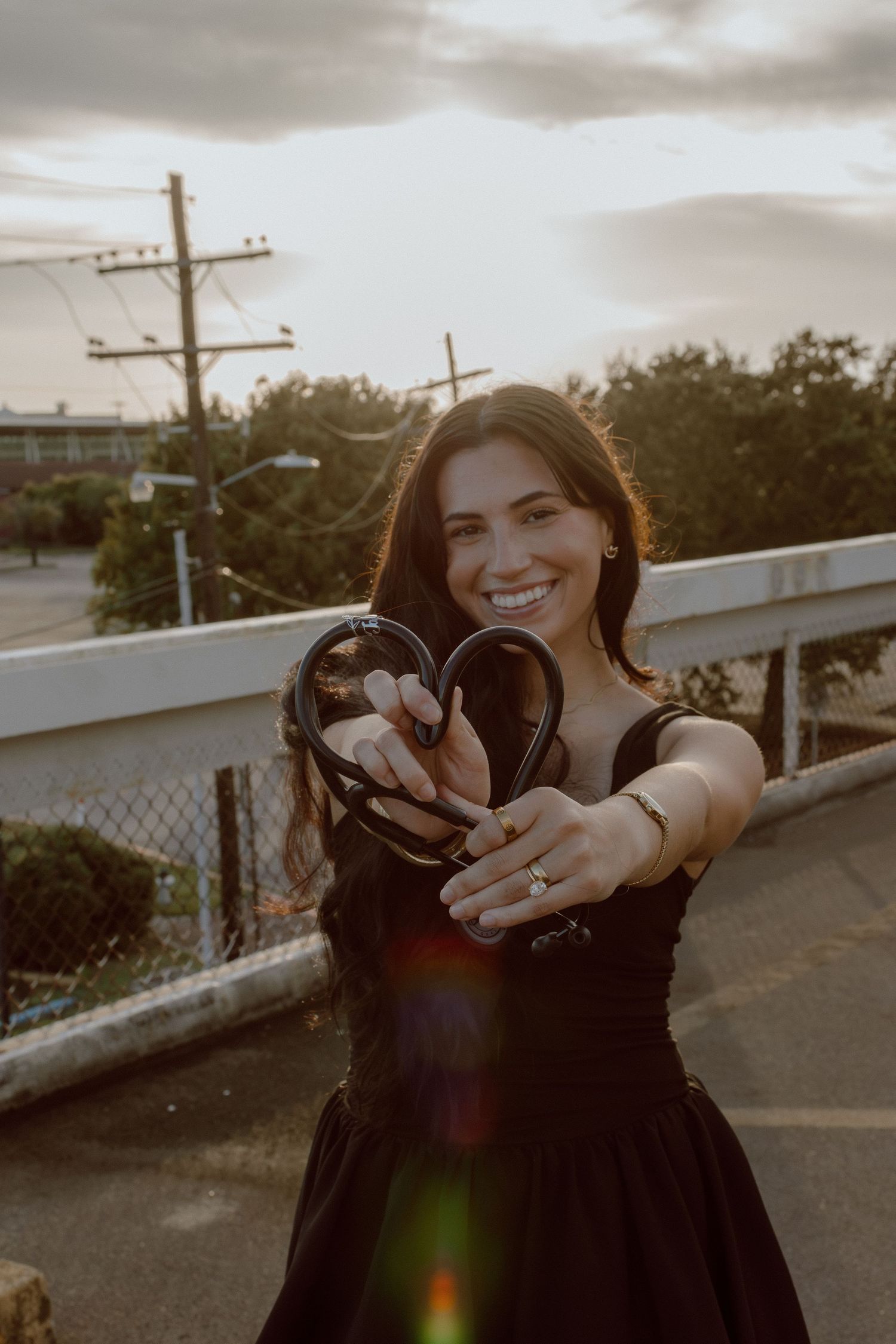 Smiling woman in black dress holding headphones in heart shape at sunset on a bridge.