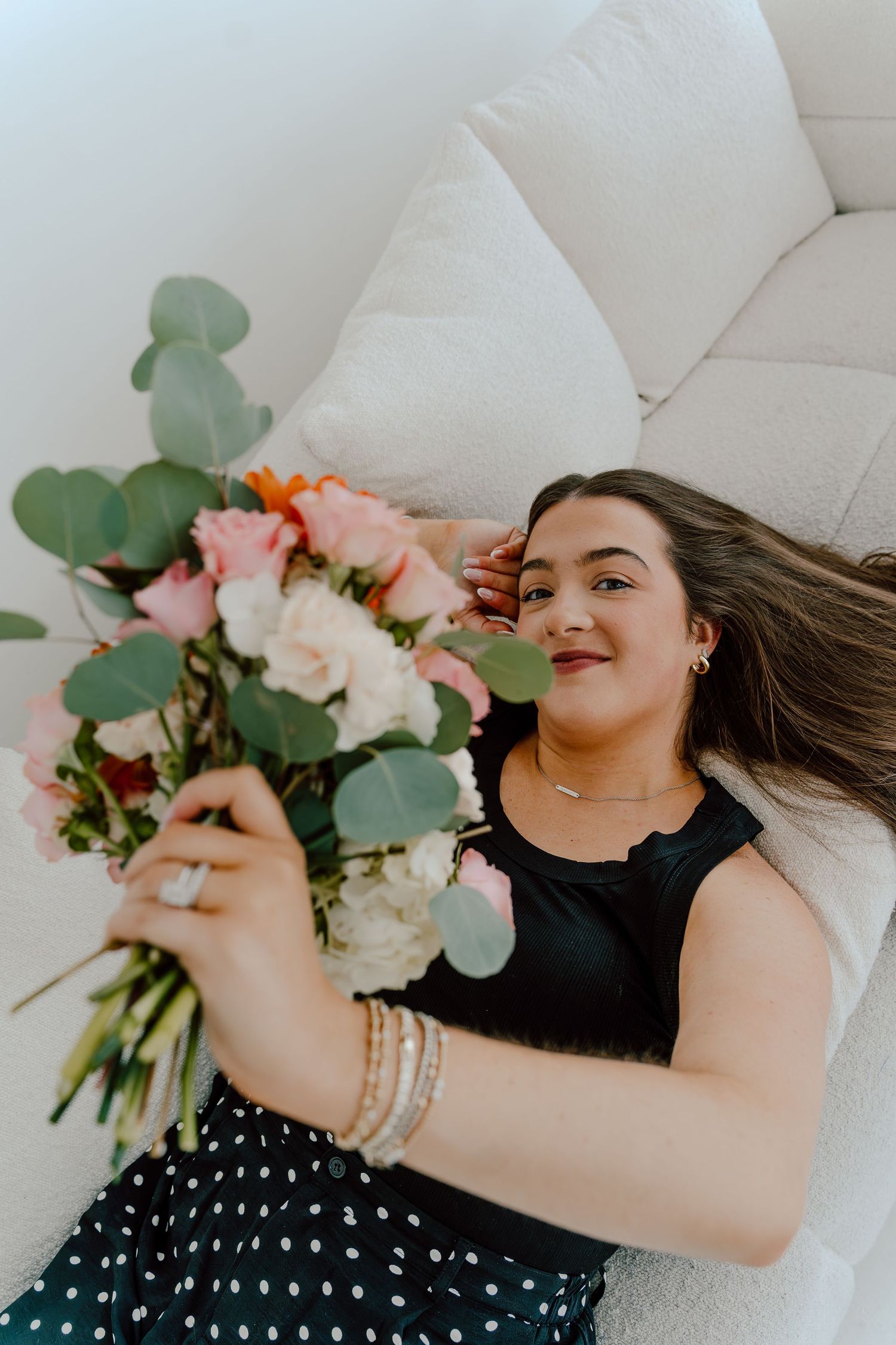 Smiling woman lying on white couch holding up a bouquet of pink and white flowers with eucalyptus.