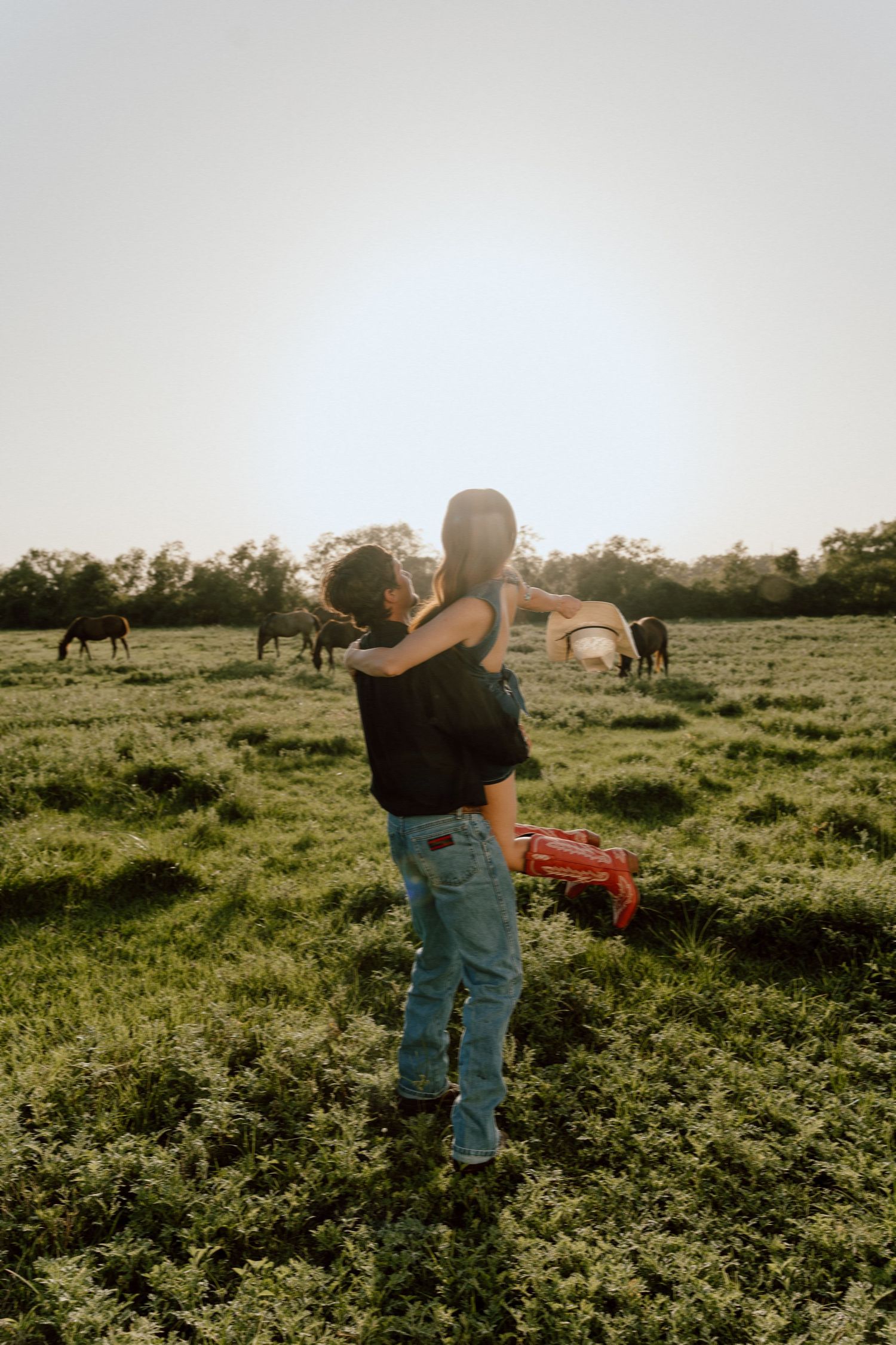 Couple embracing in a green pasture at sunset with horses grazing in the background.