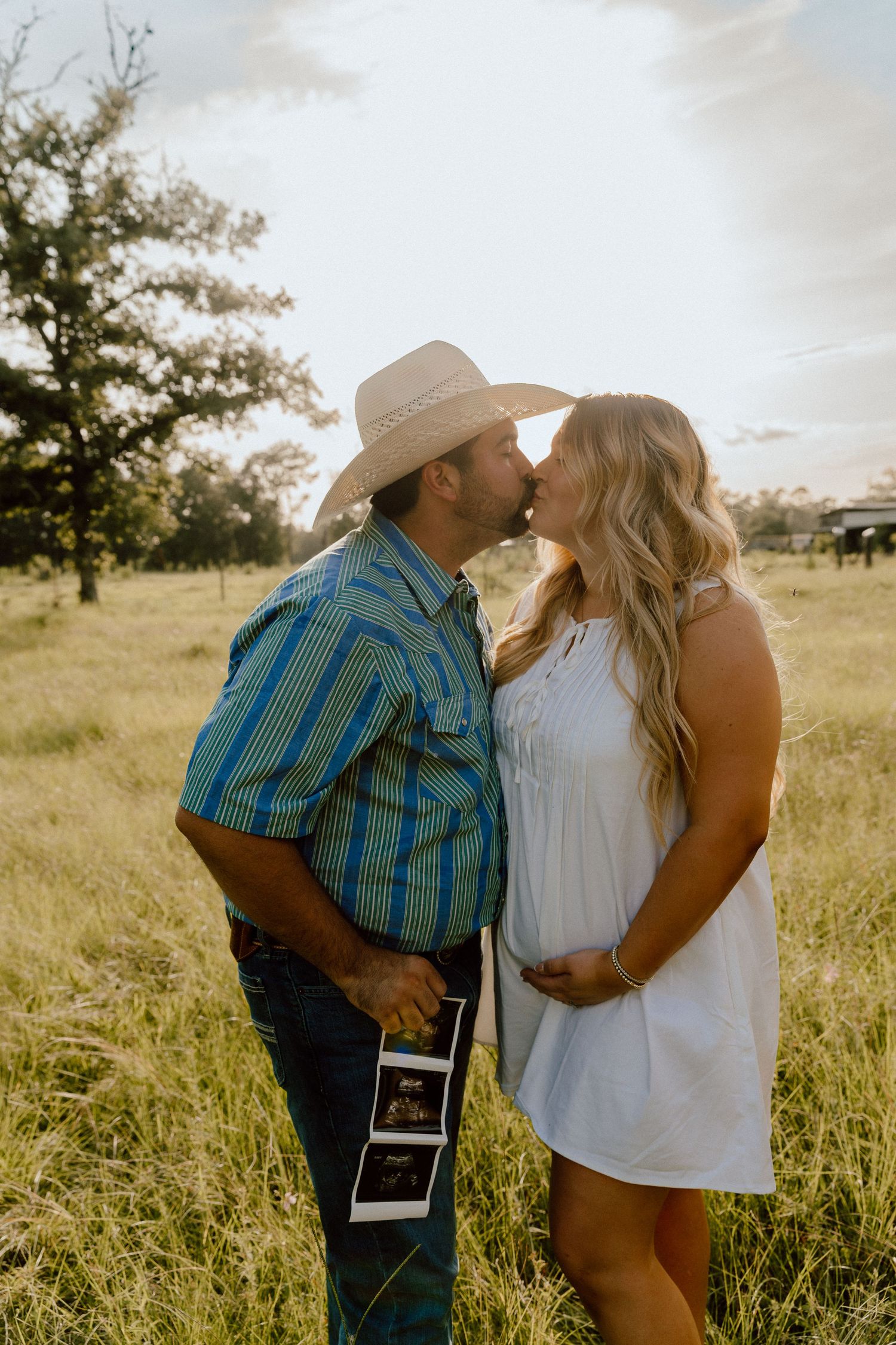 Cowboy kissing pregnant wife in white dress while holding sonogram in golden field.