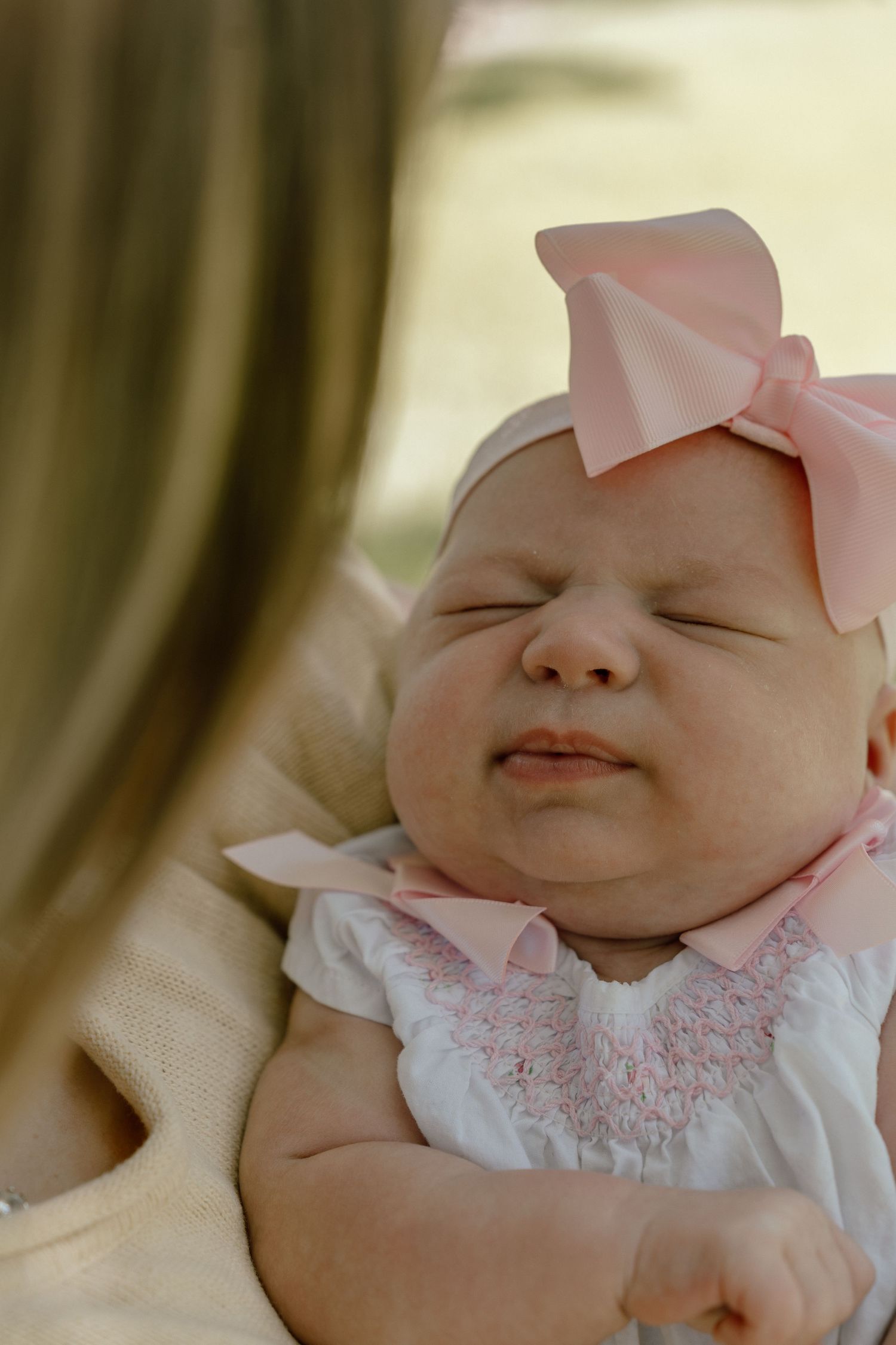 Sleeping newborn baby girl in pink smocked dress and large pink bow headband cradled in mother's arms.
