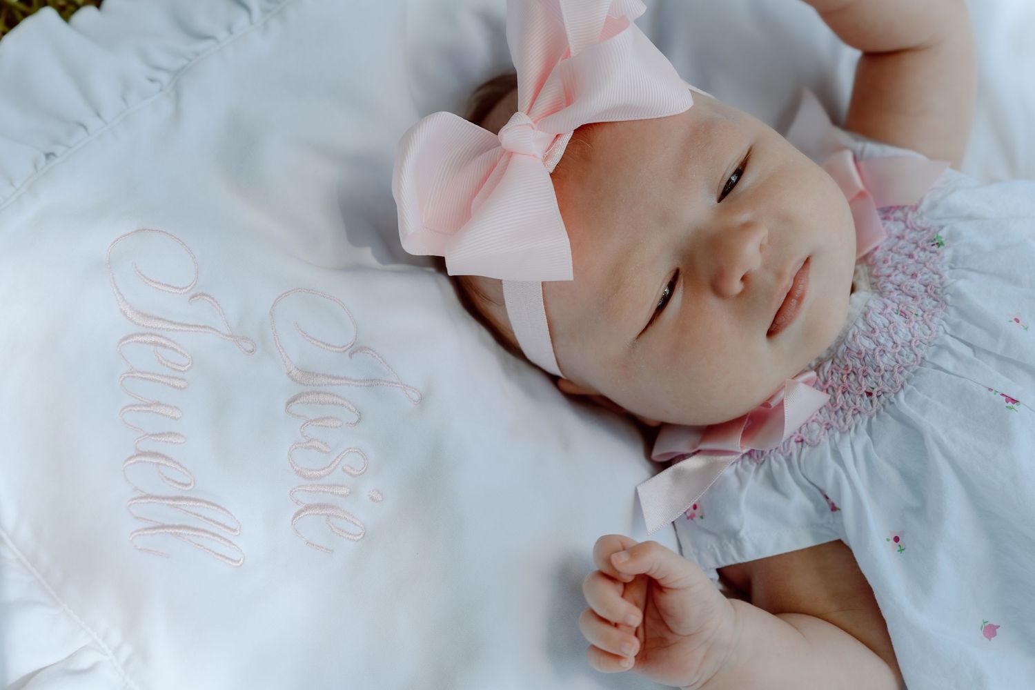 Peaceful newborn baby girl in pink bow headband lying on white embroidered blanket with eyes closed.
