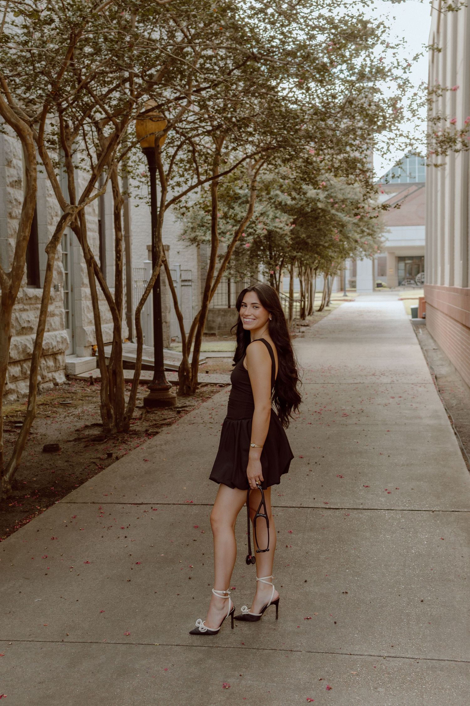Brunette woman in black dress posing on outdoor walkway with trees for graduation photos.