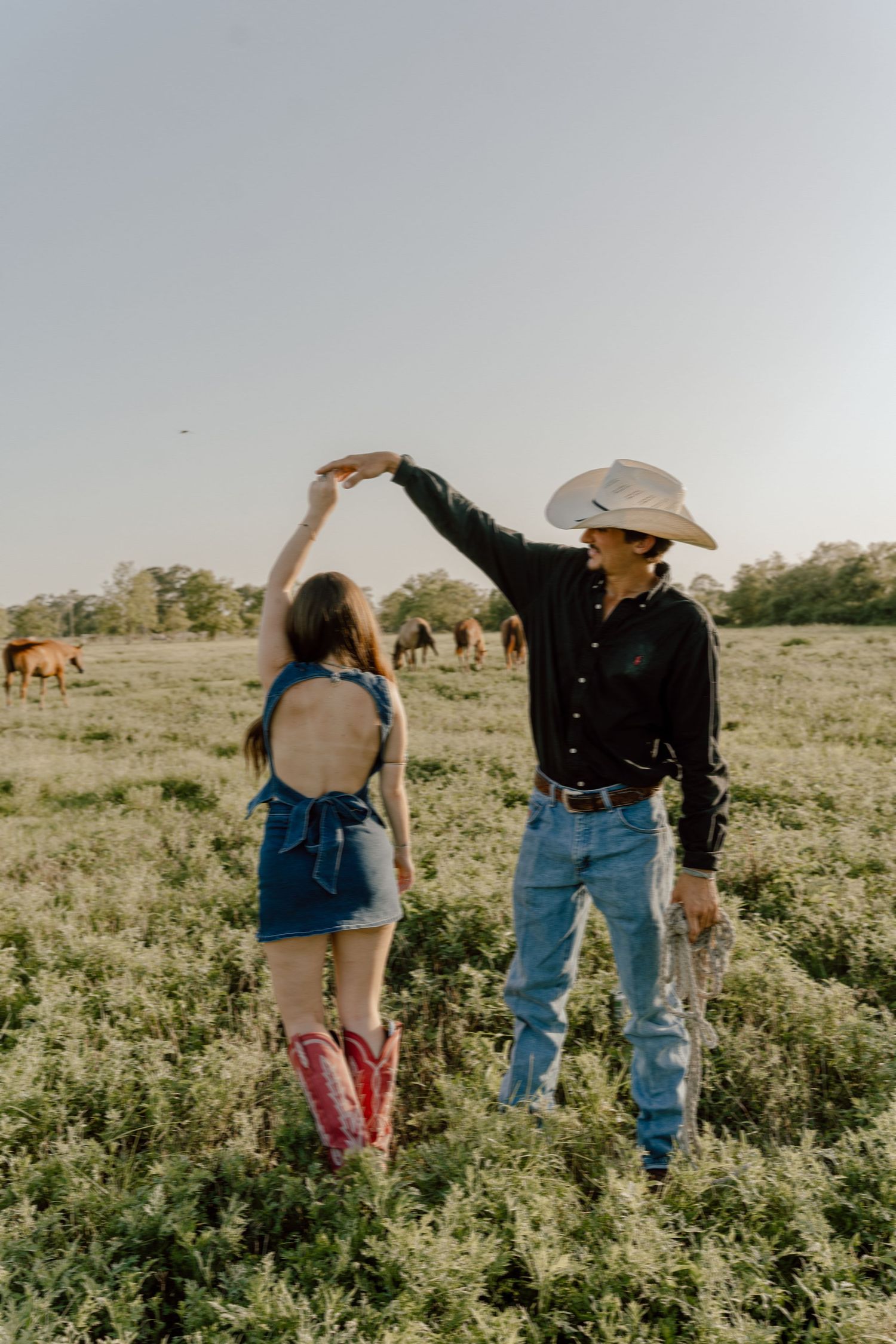 A cowboy in a white hat twirls a woman in a denim outfit and pink boots in a green field with horses.