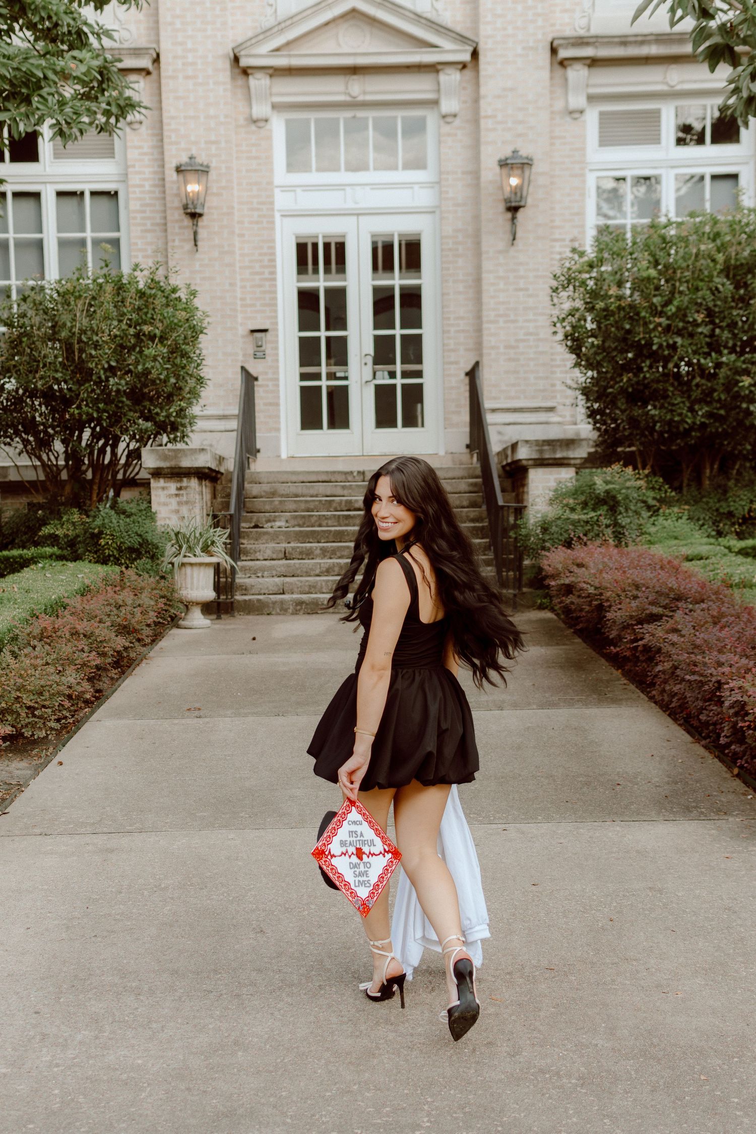 Brunette woman in black dress walking away holding graduation cap outside elegant building.