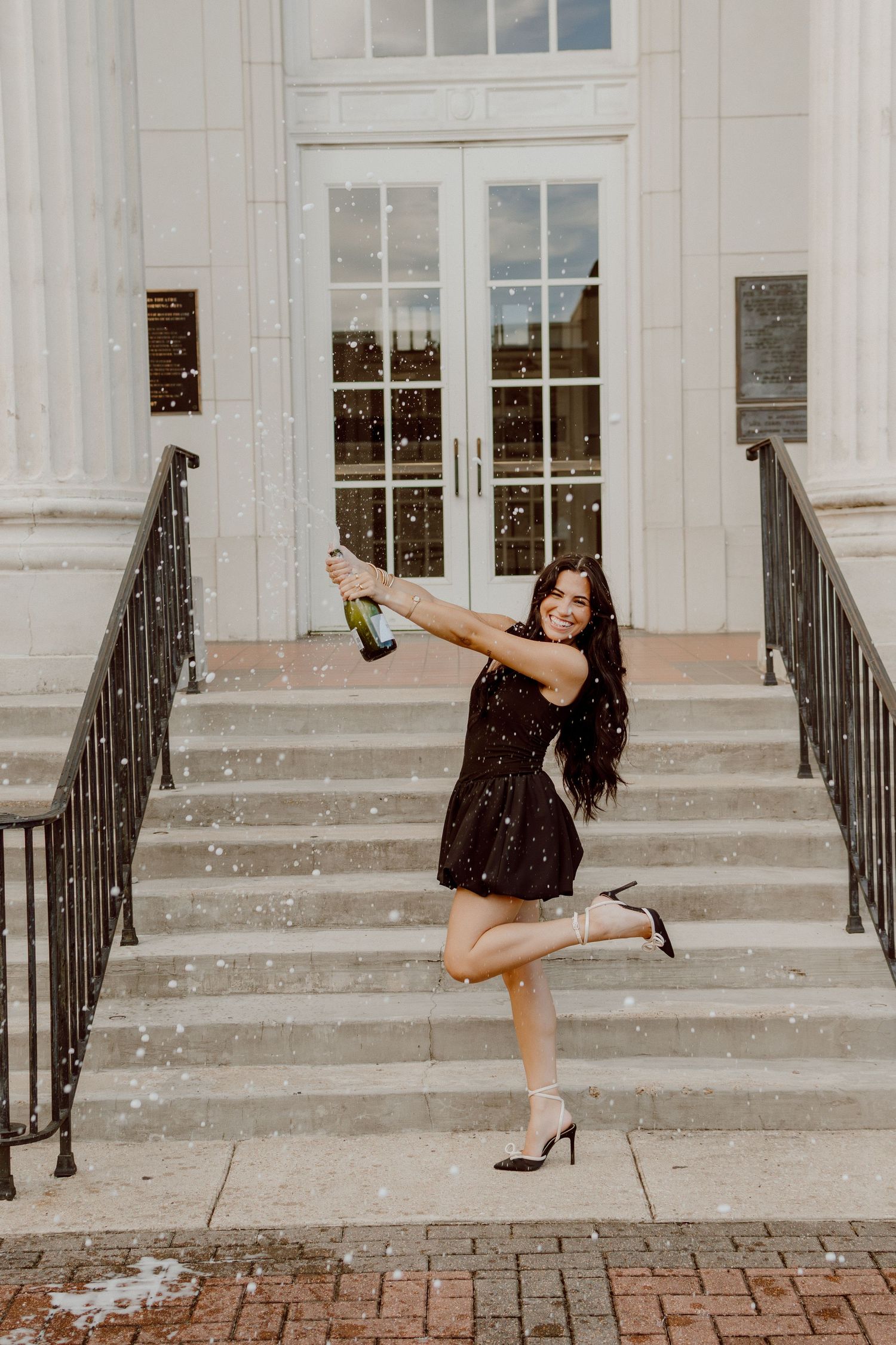 Joyful woman in black dress pops champagne bottle on steps of a classic white columned building.