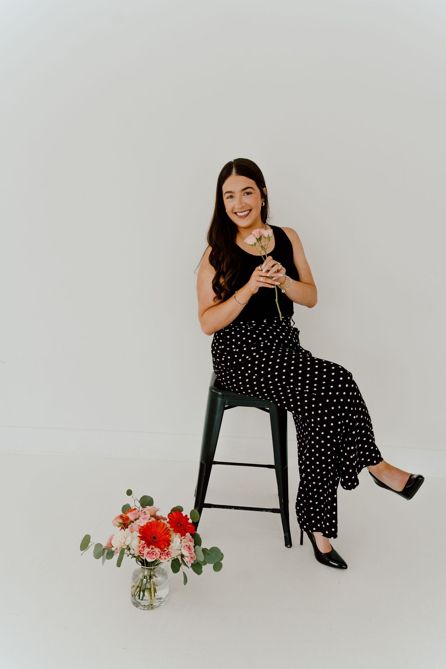 Woman in black polka dot pants sitting on stool holding a white rose with flowers on floor.