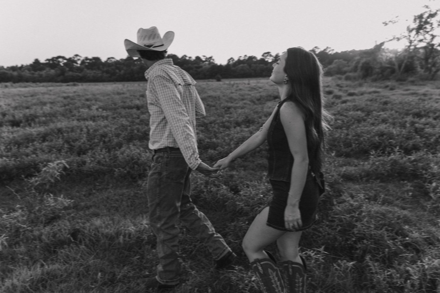 A couple holds hands in a black and white photo while walking through a lush open field at dusk.