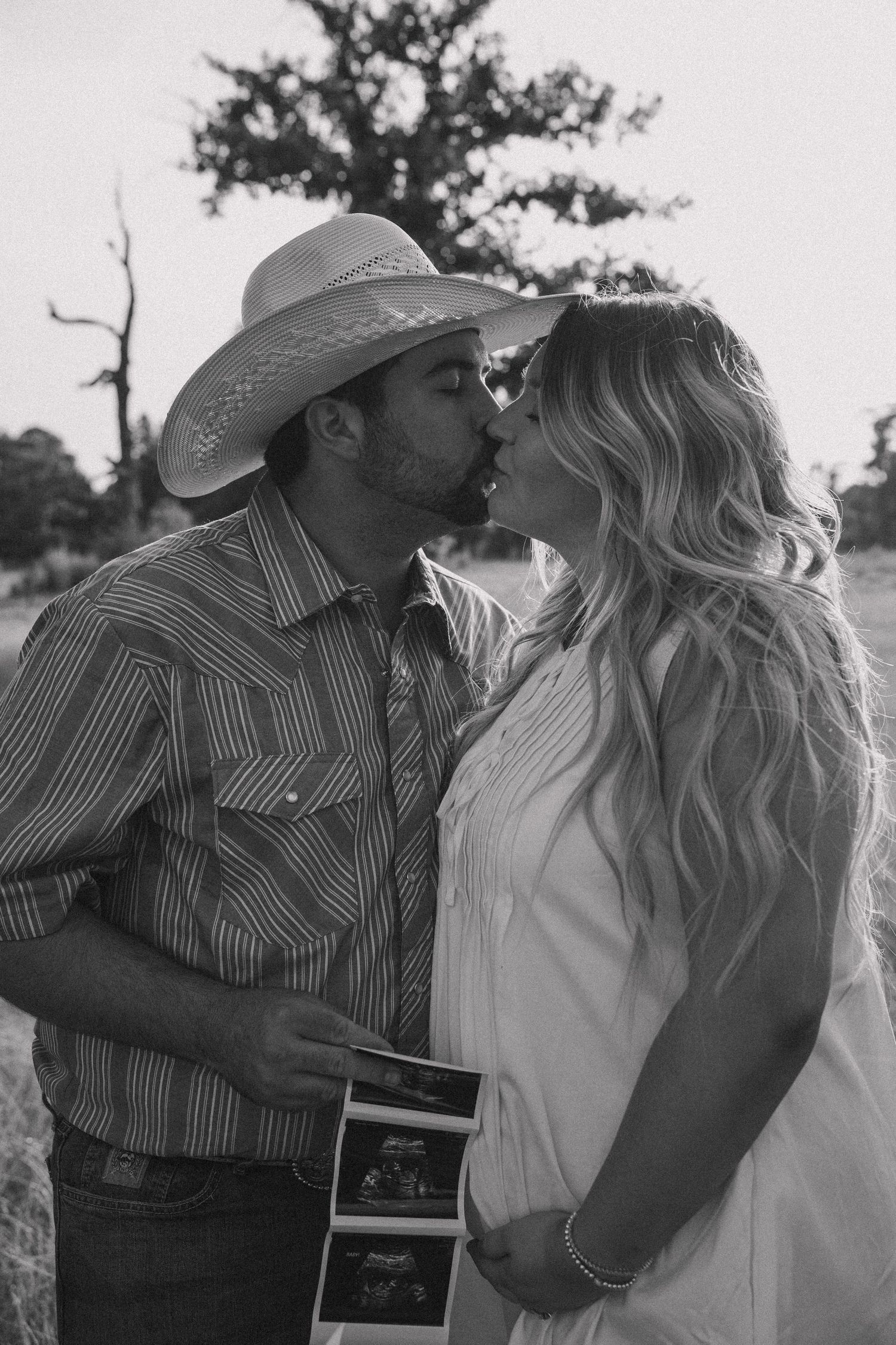 Black and white photo of expecting couple kissing while holding baby sonogram in field.