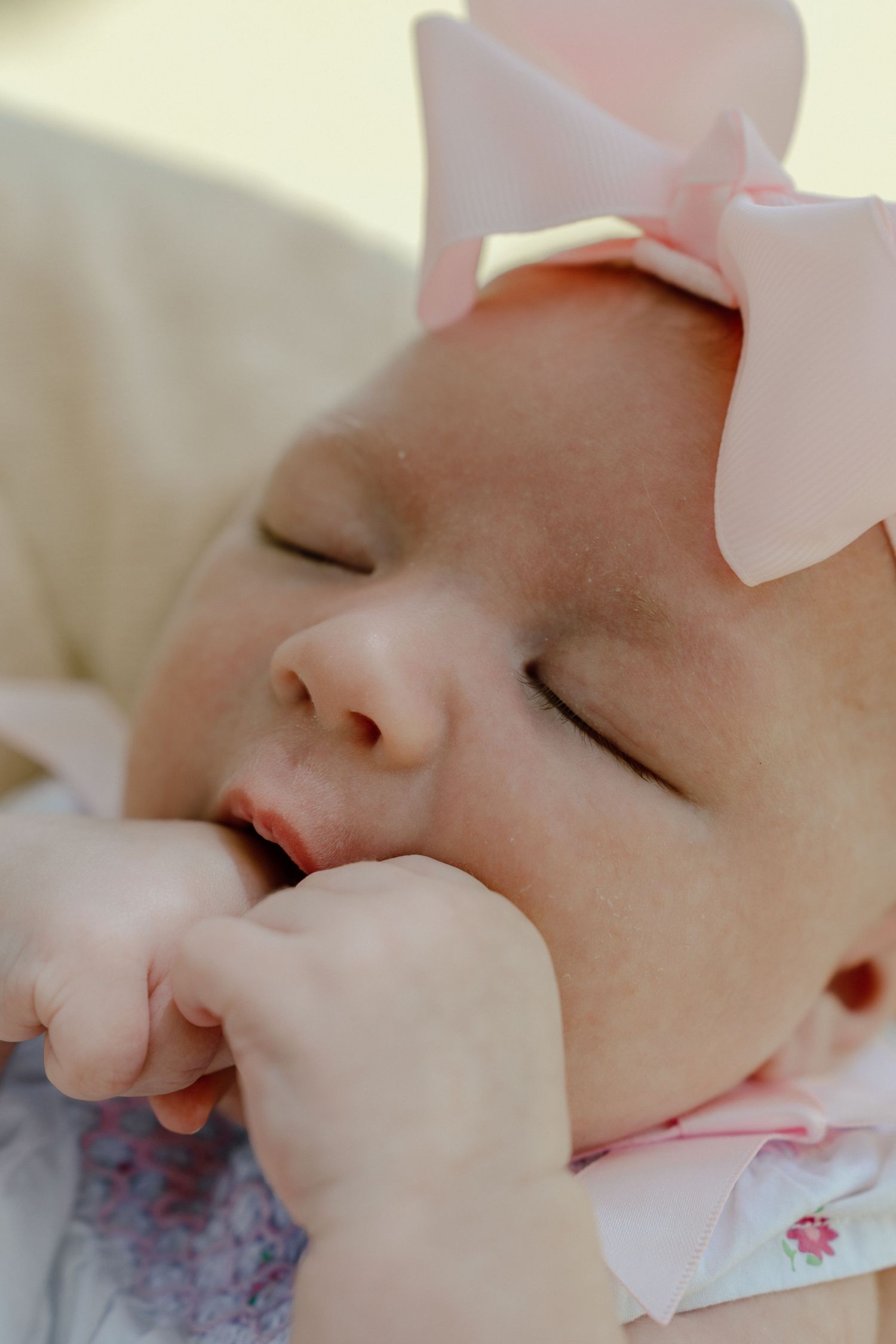 Close-up of sleeping newborn baby girl with pink bow headband sucking her fist in soft natural light.