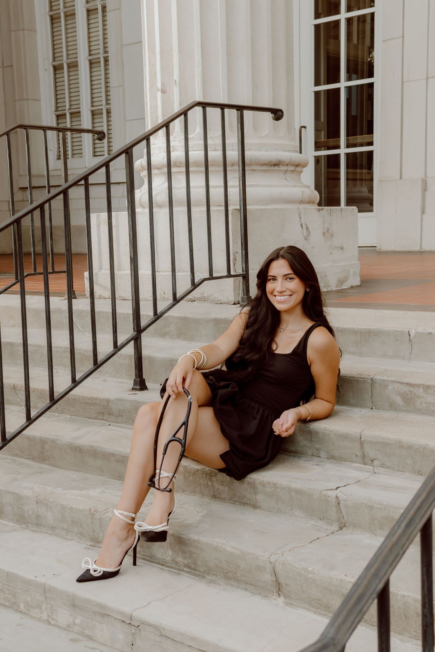 Smiling brunette woman in black dress sitting on marble steps with heels for senior photos.