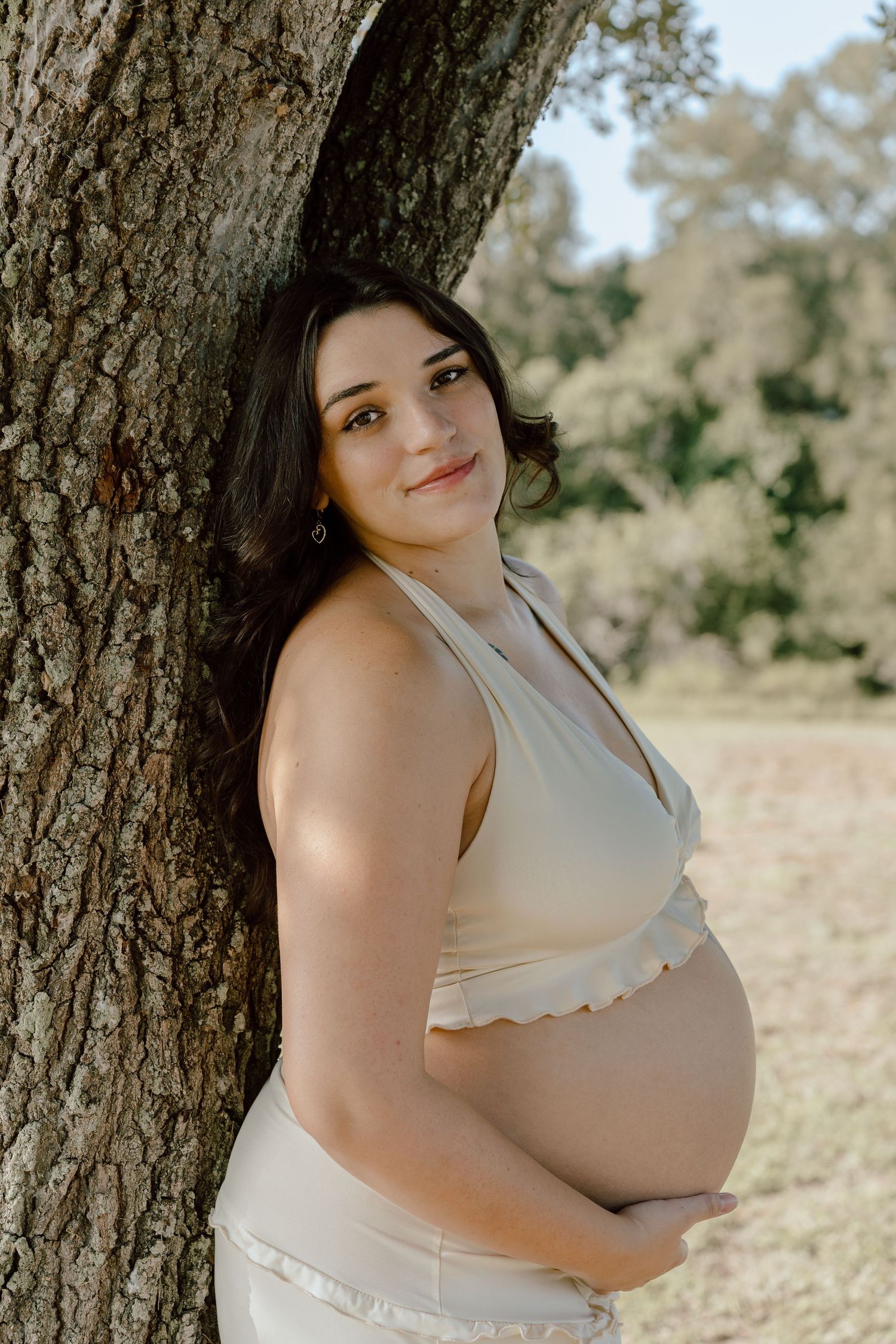 Smiling pregnant woman in a cream halter top leaning against a large tree outdoors.