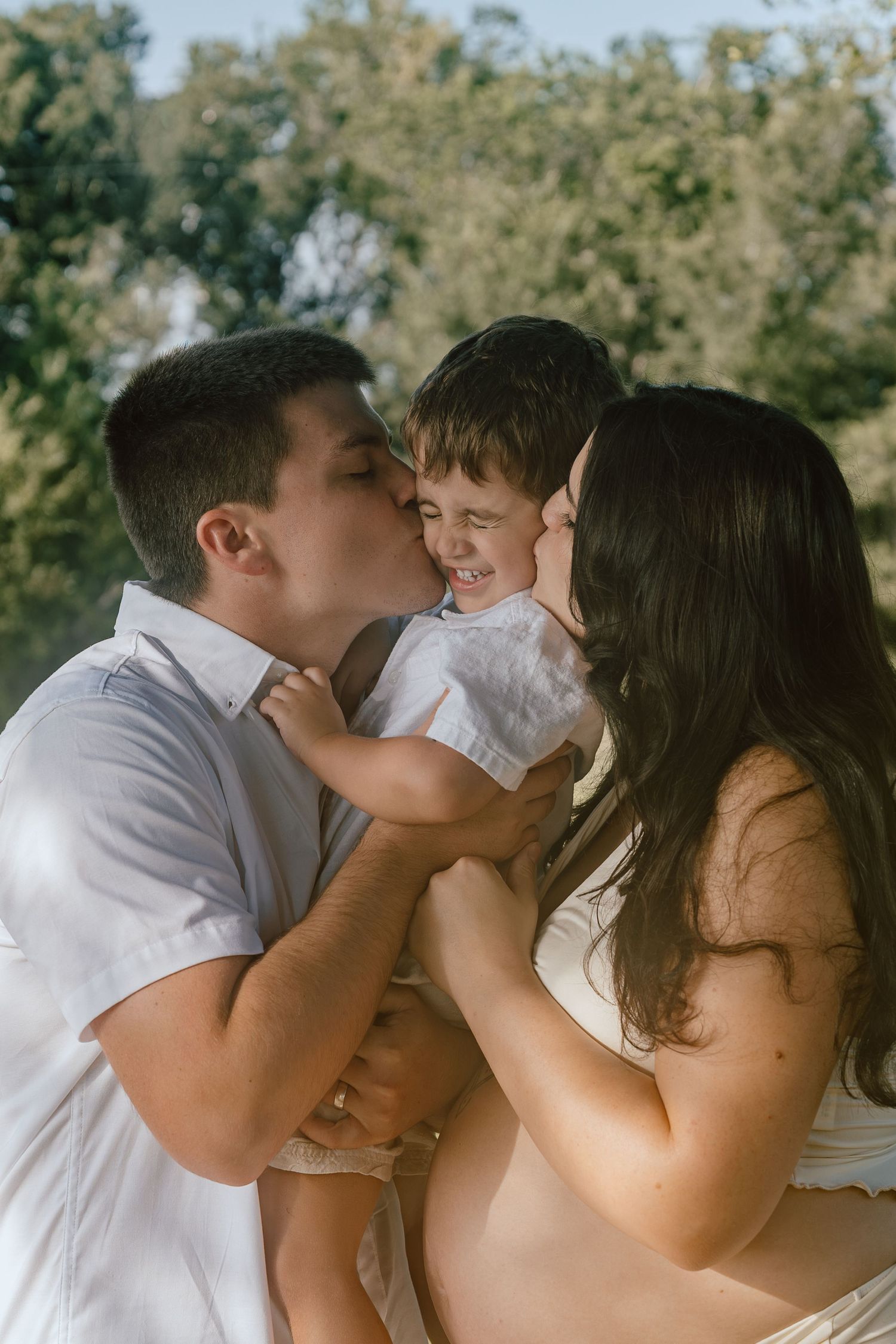 Parents kissing smiling toddler son on cheeks during outdoor maternity photo session.