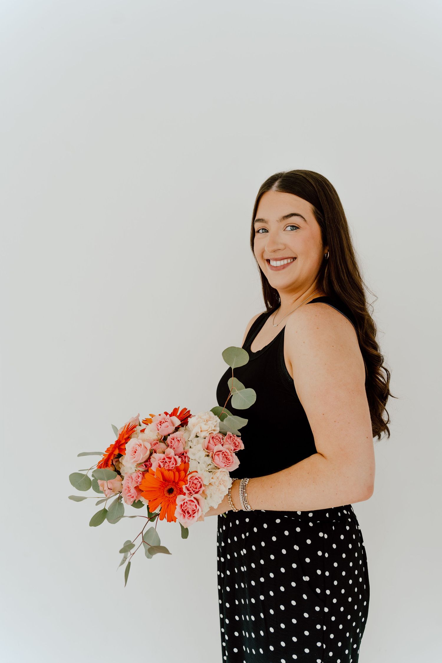 Smiling woman in black tank top and polka dot skirt holding a colorful bouquet against white background.