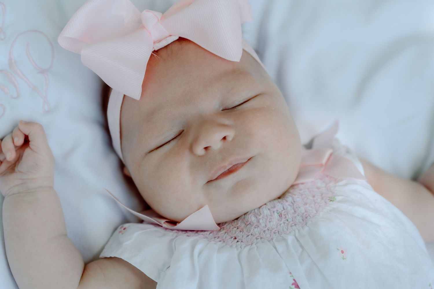Serene newborn baby girl in pink smocked outfit and bow headband sleeping peacefully on white blanket.