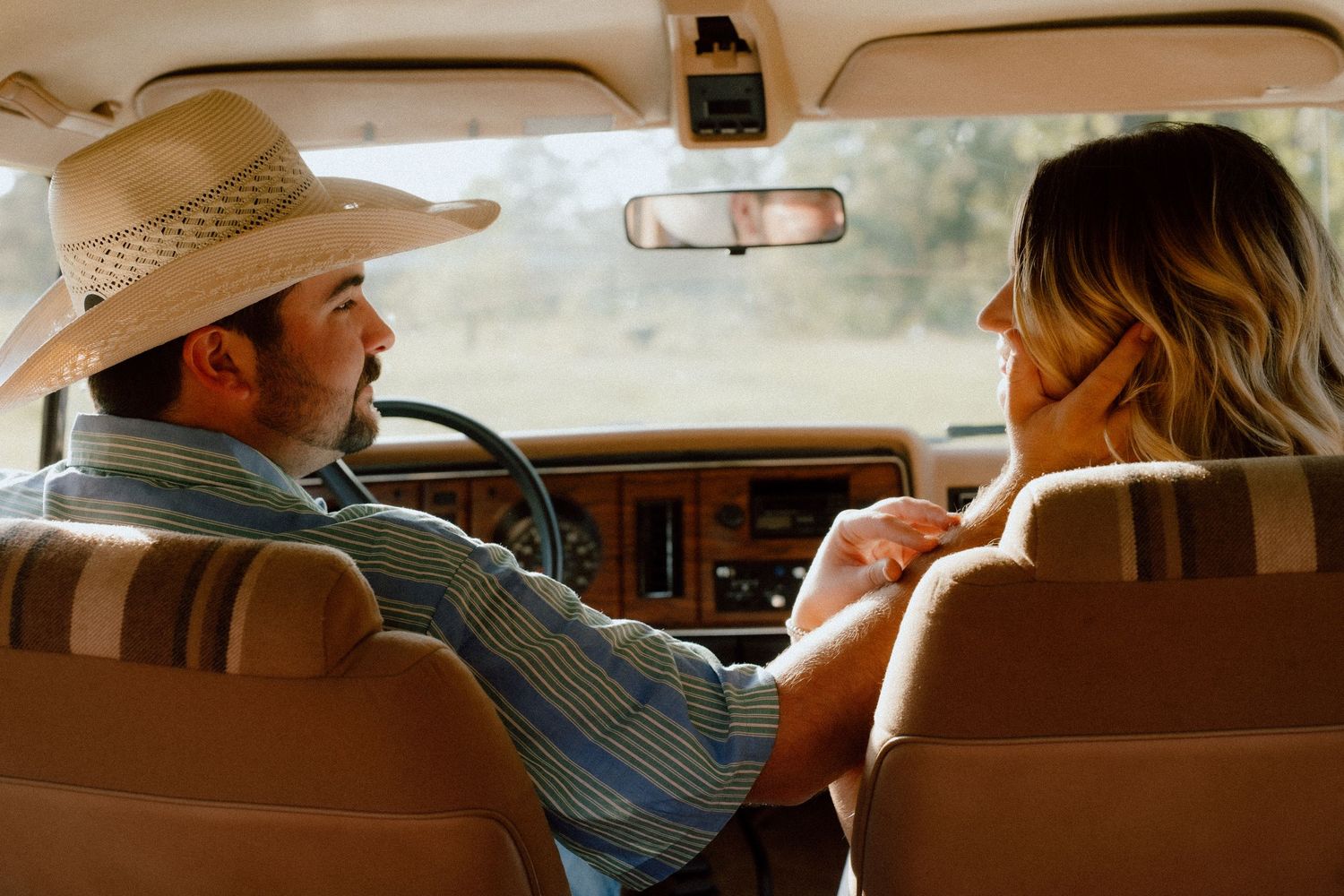 A man in a cowboy hat reaches toward a woman in a vintage car interior, sharing a tender moment.