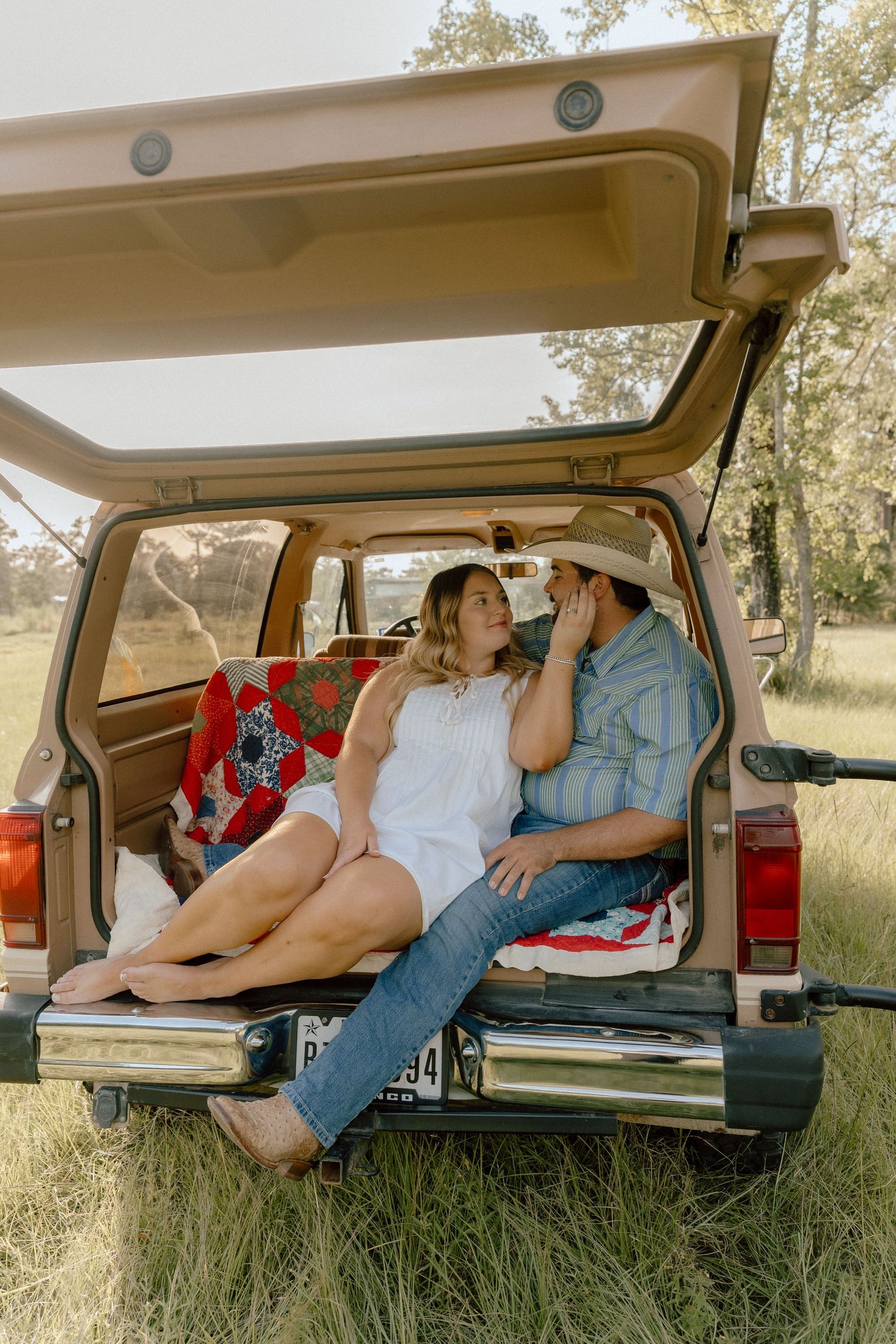 Couple sitting in the open trunk of a vintage SUV in a grassy field at sunset.