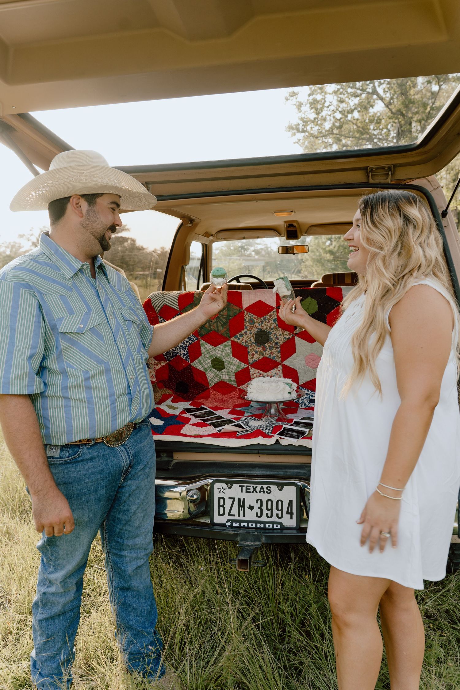 Couple standing at open tailgate of vintage Ford Bronco with Texas plates and quilt backdrop.