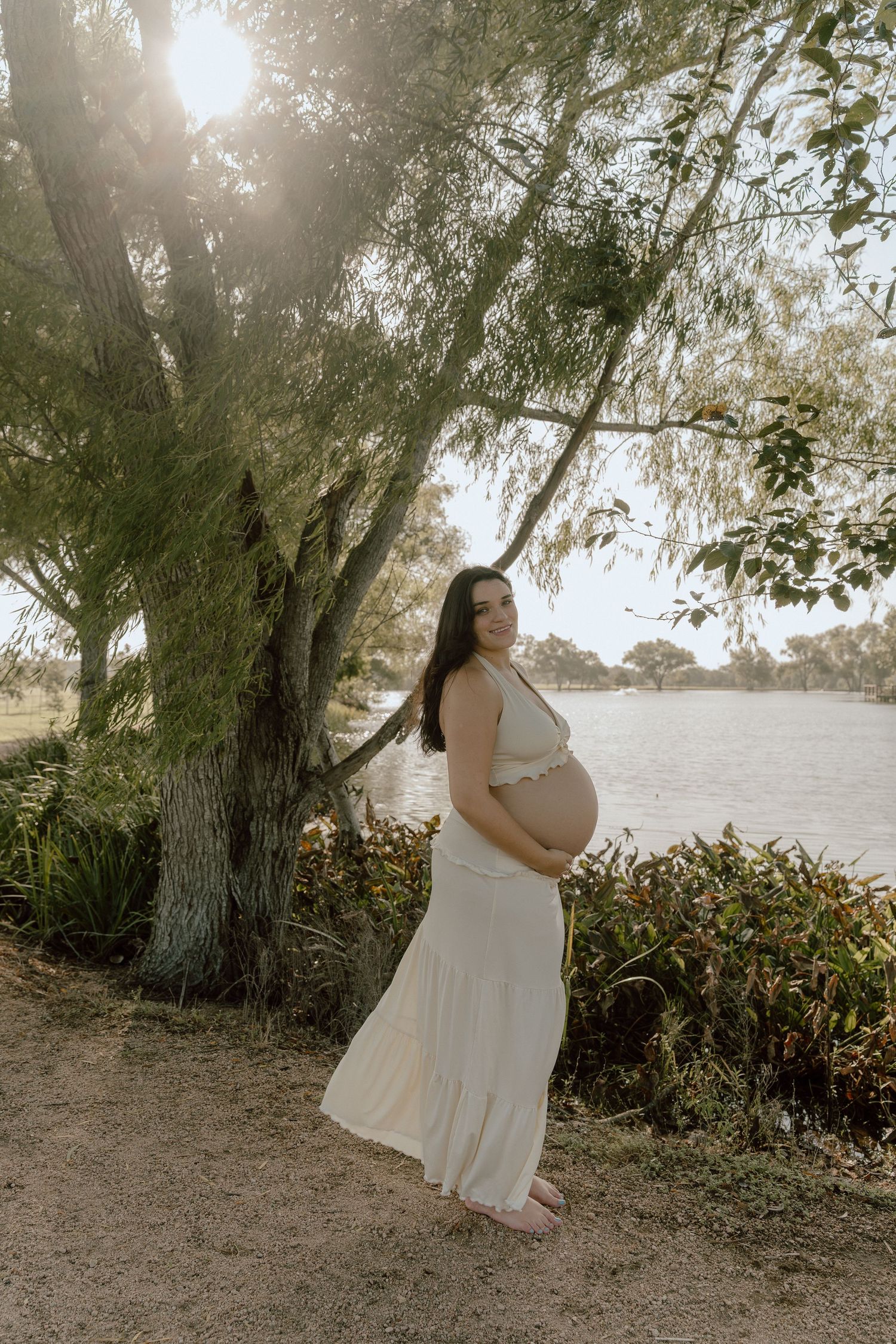 Pregnant woman in a flowing cream dress standing by a sunlit willow tree near water.
