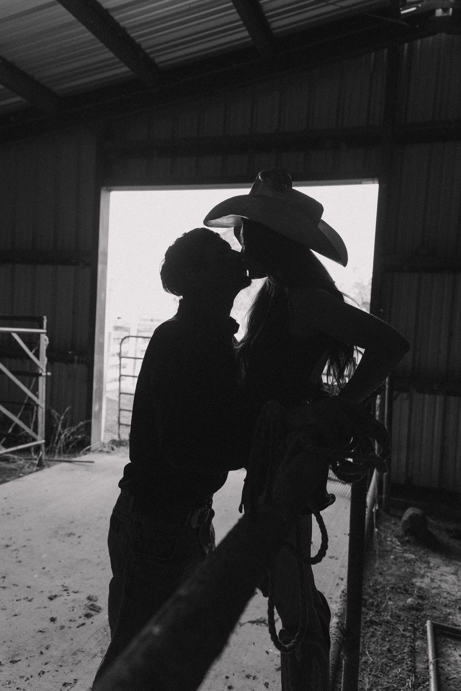 A couple shares a romantic kiss silhouetted in a barn doorway in a dramatic black and white photograph.