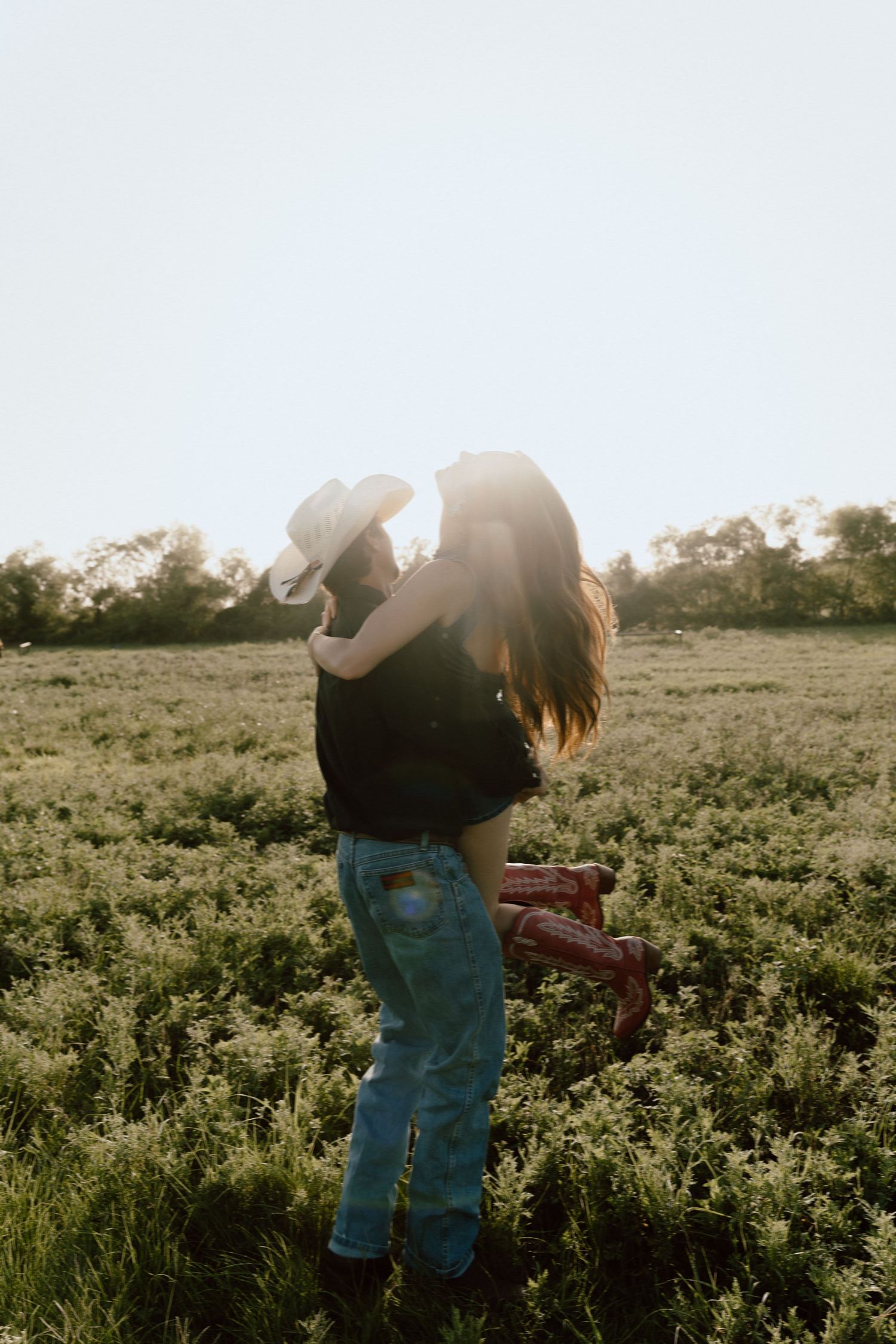 Man lifting woman in a sunny green field at golden hour, woman wearing red cowboy boots.