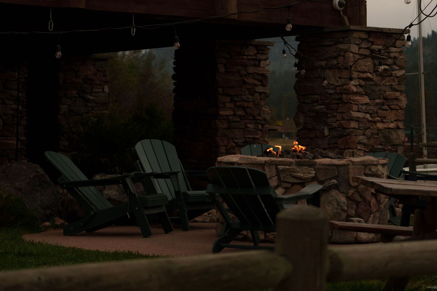 Adirondack chairs gathered around a stone outdoor fireplace glowing at dusk on a patio.