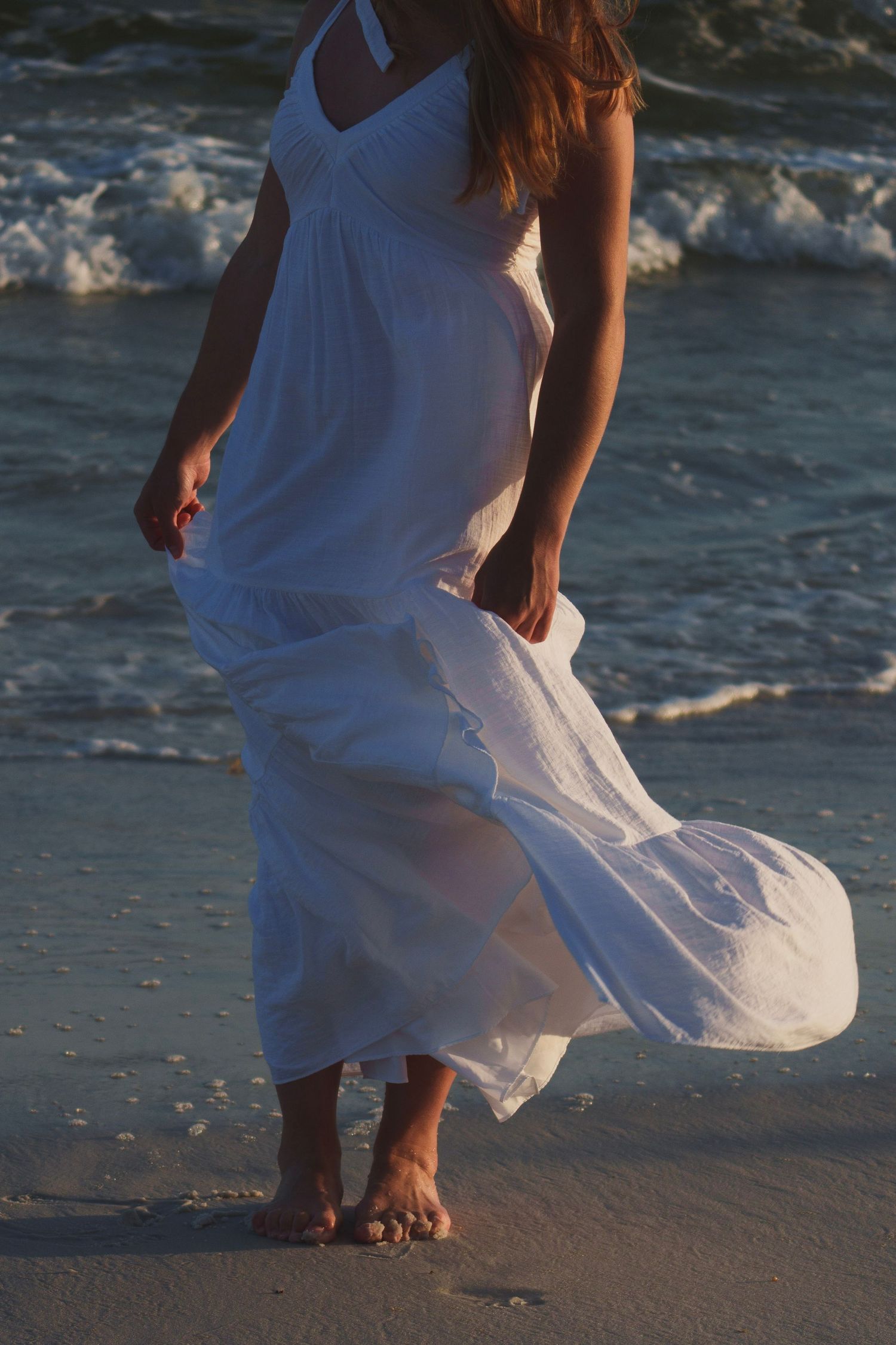 Woman in flowing white maxi dress walking barefoot on a sandy beach at sunset with waves crashing.