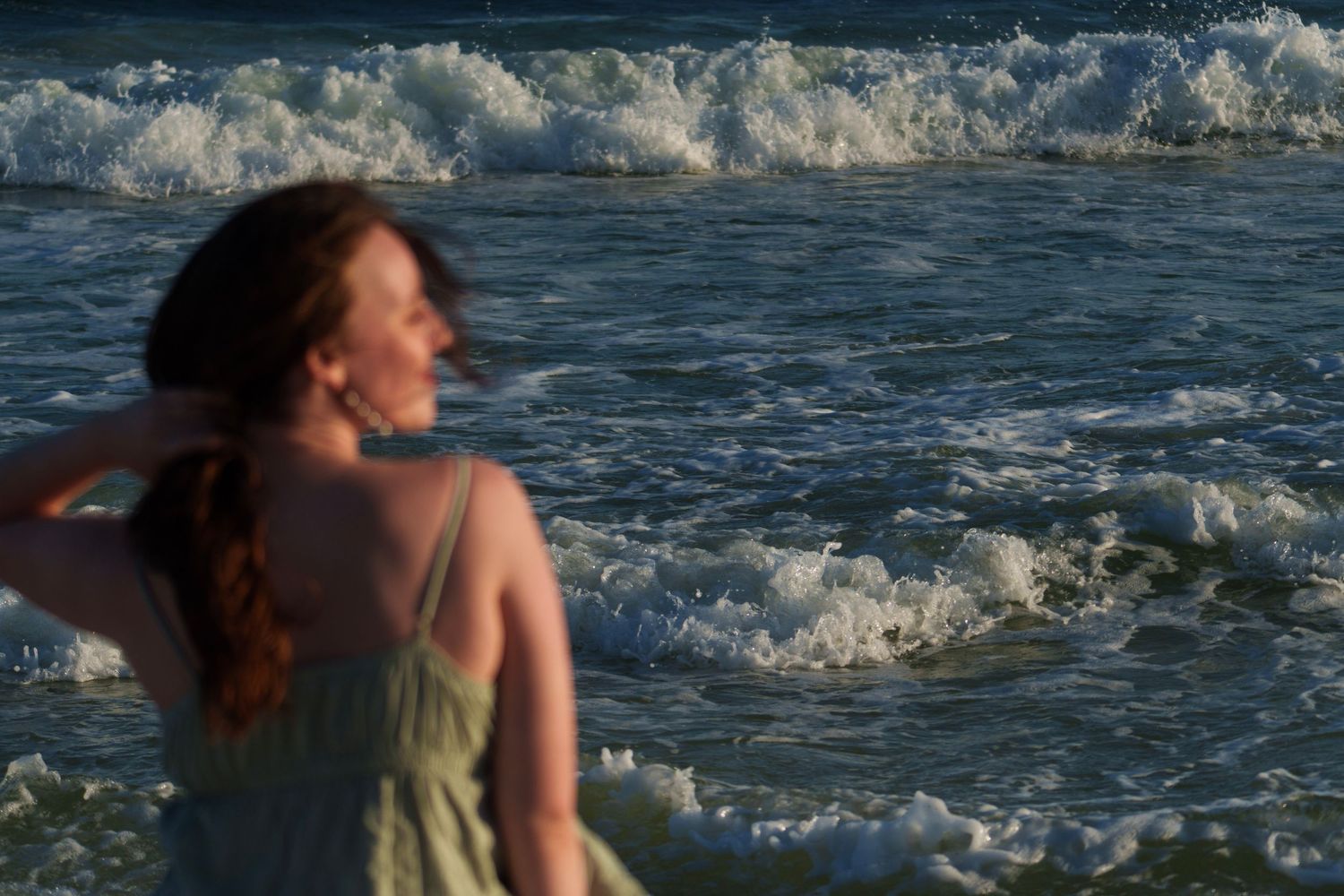 Red-haired woman in green dress standing near crashing ocean waves at golden hour sunset.