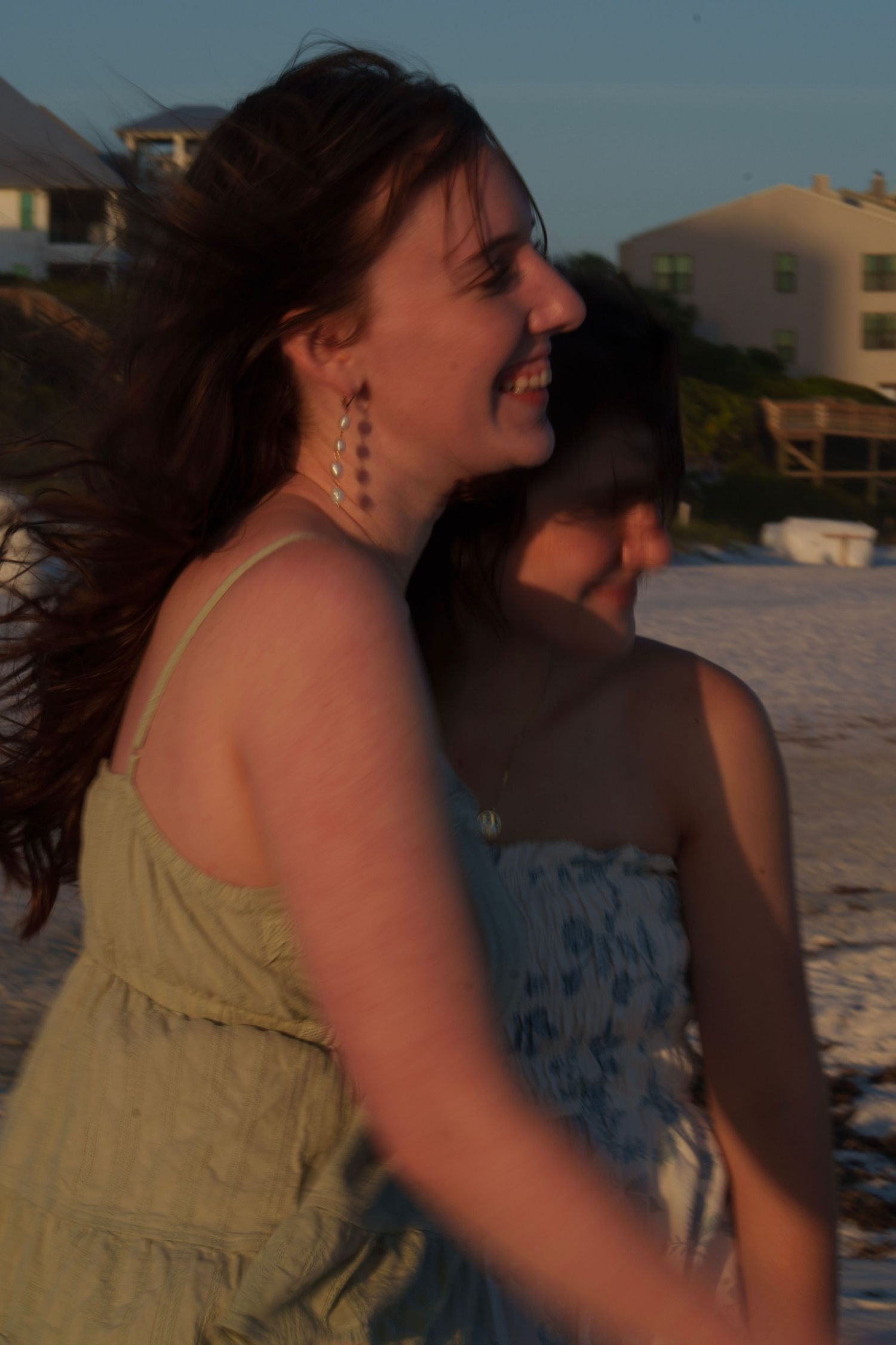 Two laughing young women in summer dresses enjoying a breezy sunset on a sandy beach.