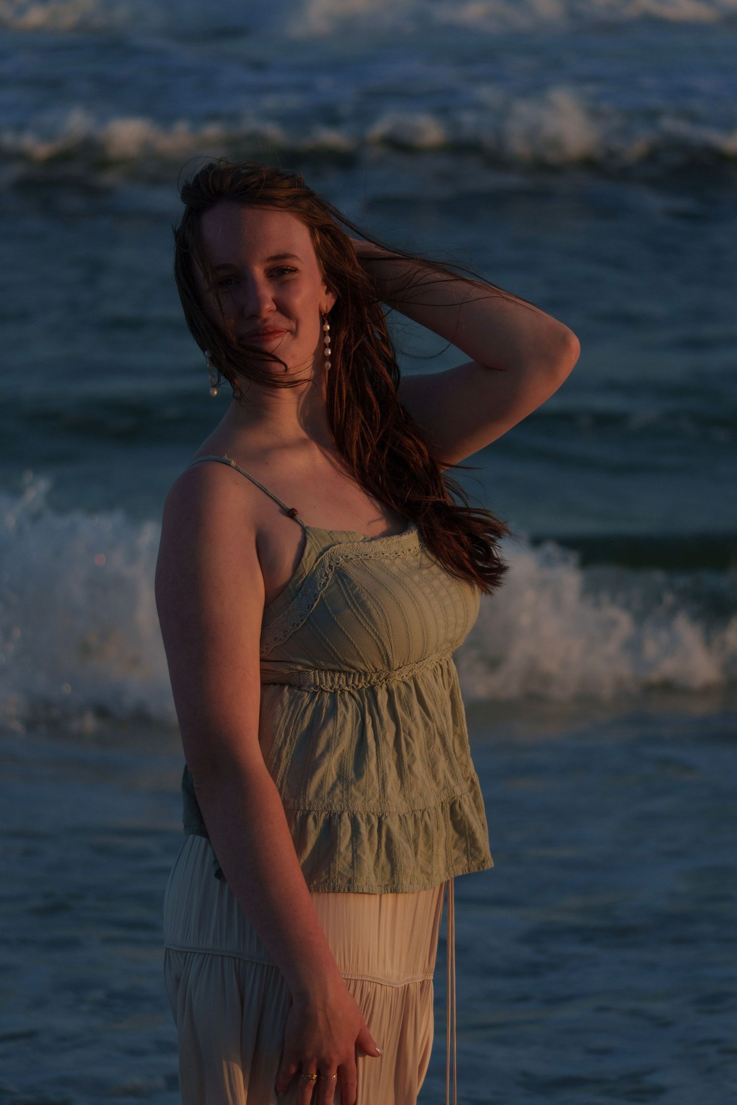 Young woman in sage green top and white skirt posing on beach with dramatic ocean waves at dusk.