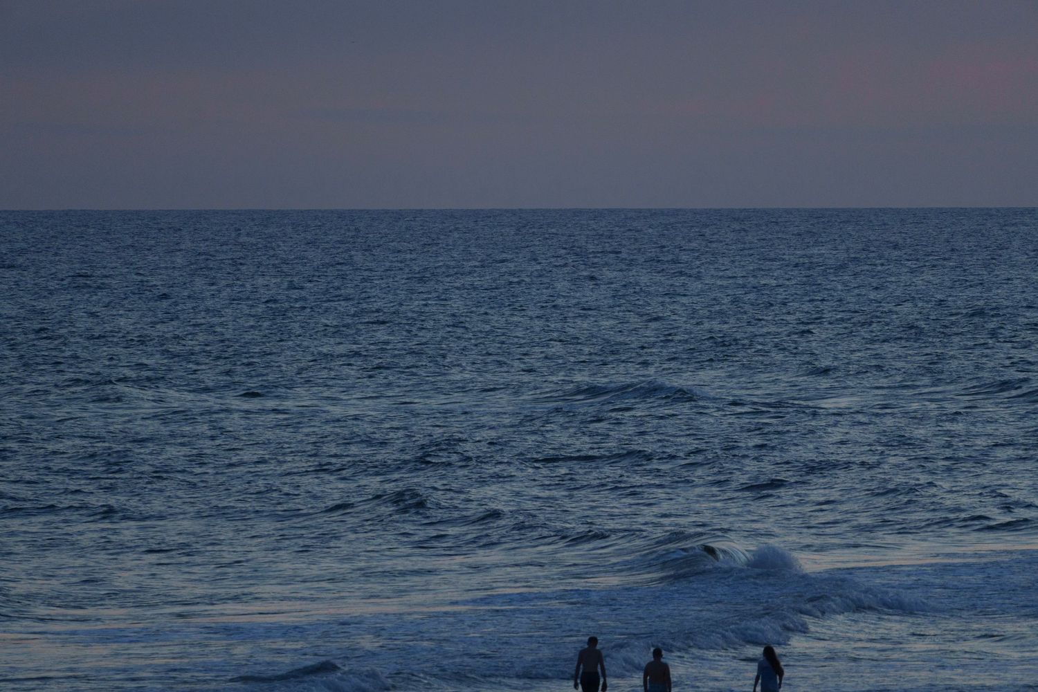 Three silhouetted figures stand at the ocean shoreline during a blue dusk twilight.
