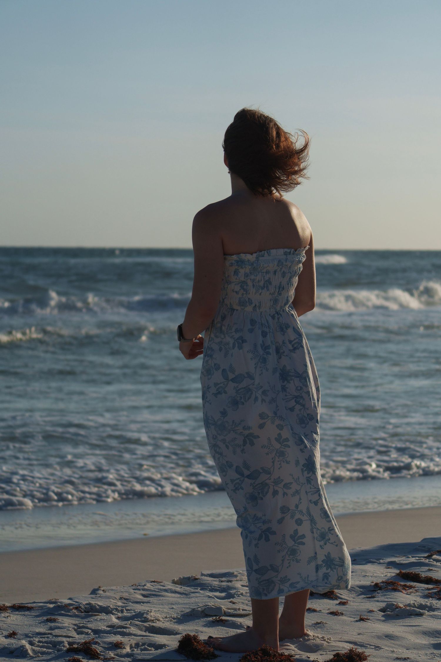 Woman in blue floral strapless maxi dress standing on sandy beach gazing at ocean waves at dusk.