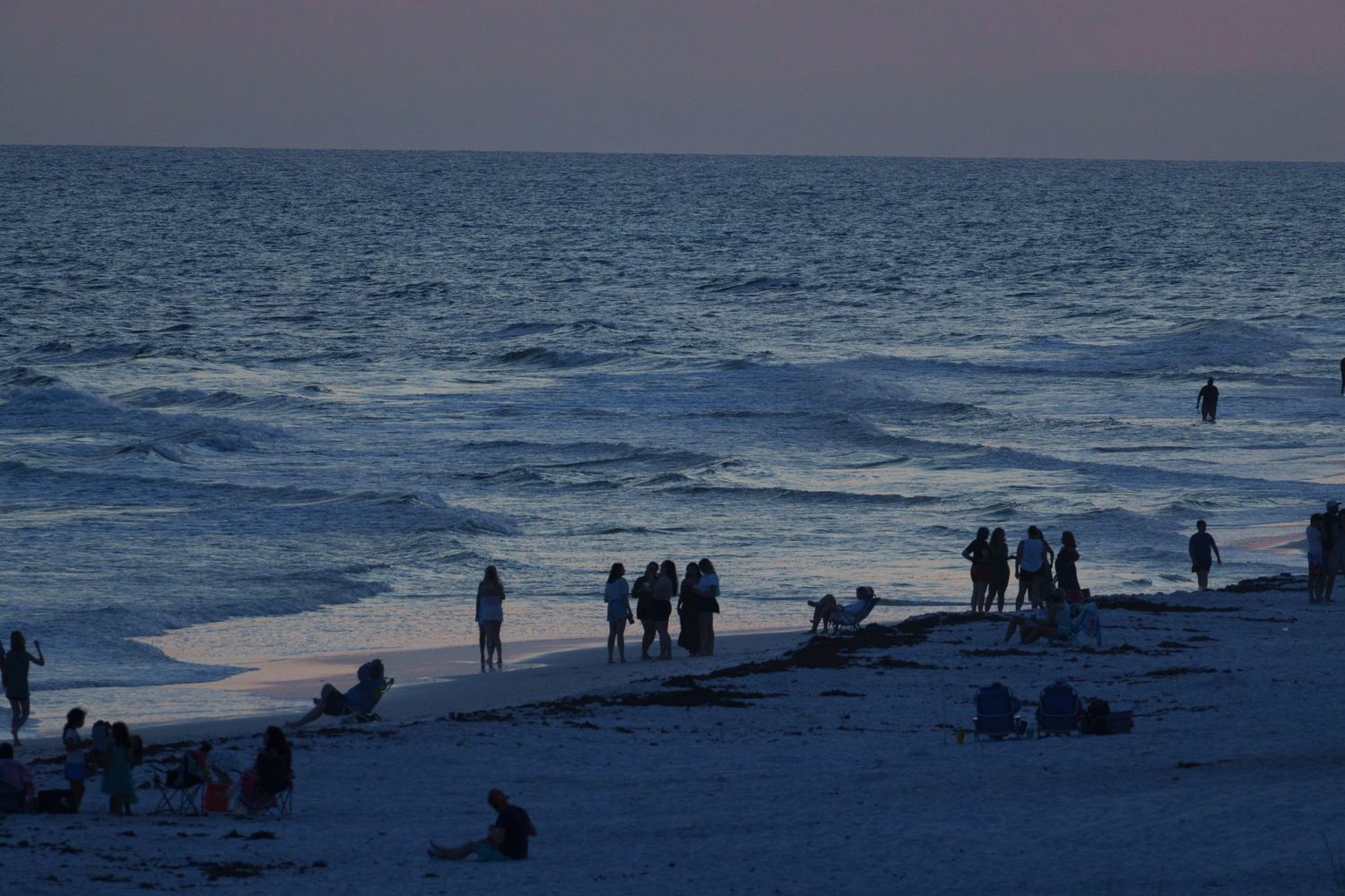 Beachgoers gather along a sandy shore at sunset with gentle ocean waves in the background.
