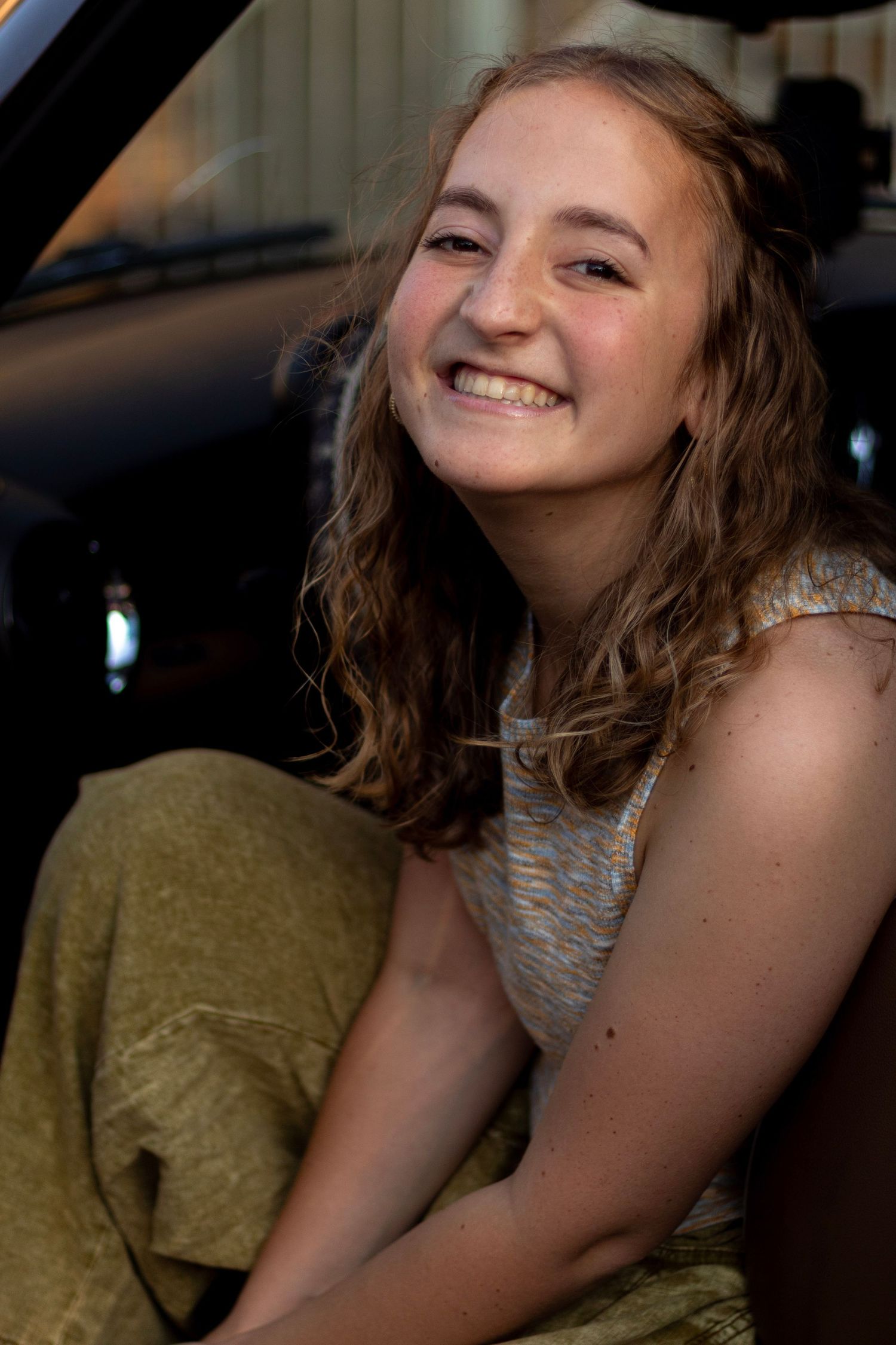 Smiling teenage girl with wavy hair sitting in a car doorway in warm sunlight.
