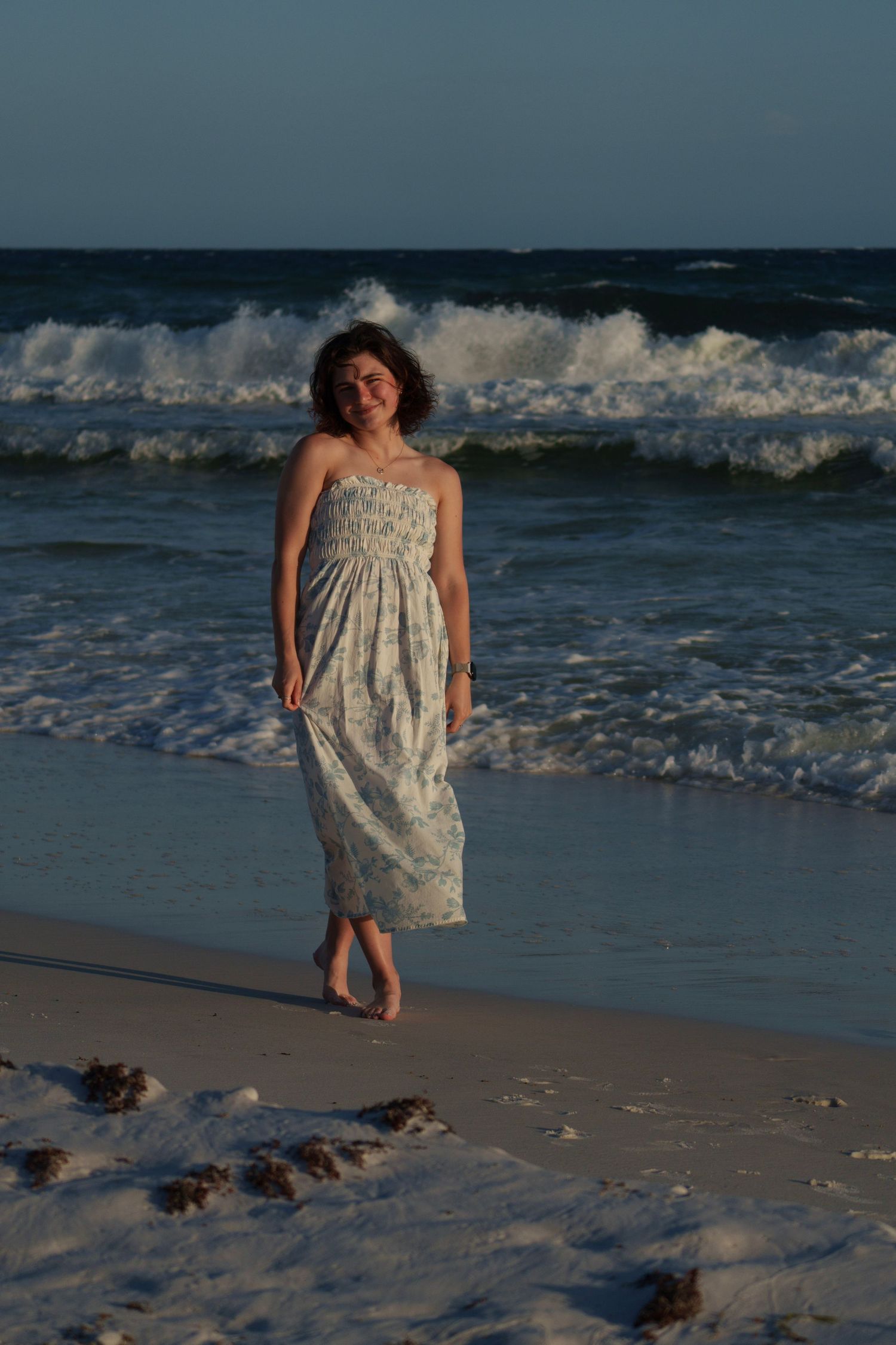 Woman in floral strapless maxi dress walking barefoot on sandy beach at sunset.