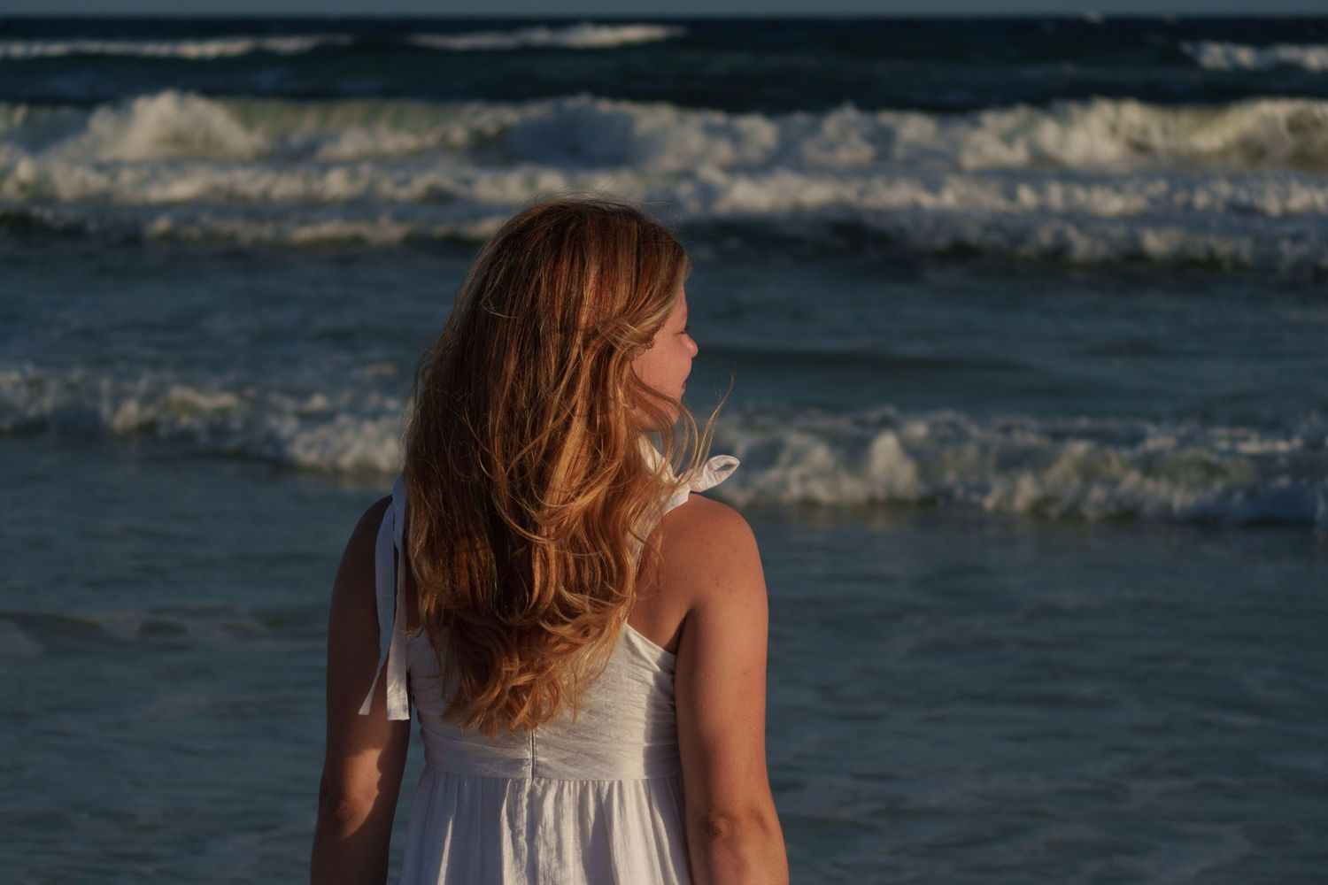 Young woman in white dress standing on sandy beach watching ocean waves at sunset.