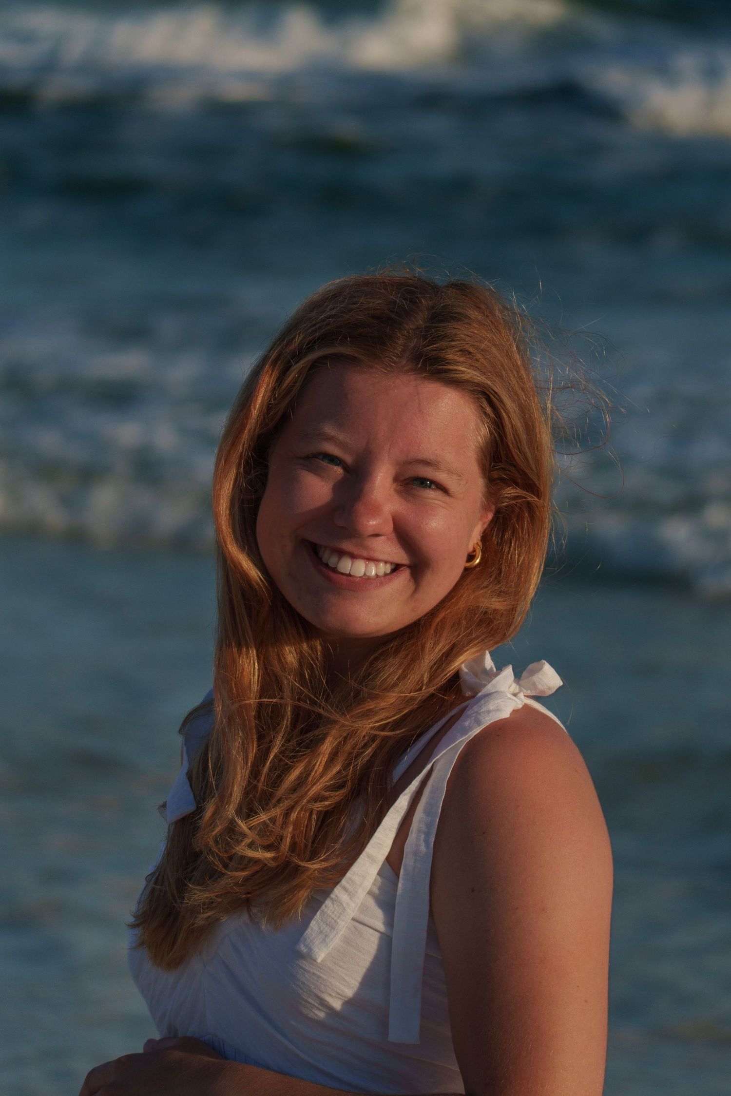 Smiling woman with long hair in white top at golden hour beach sunset.