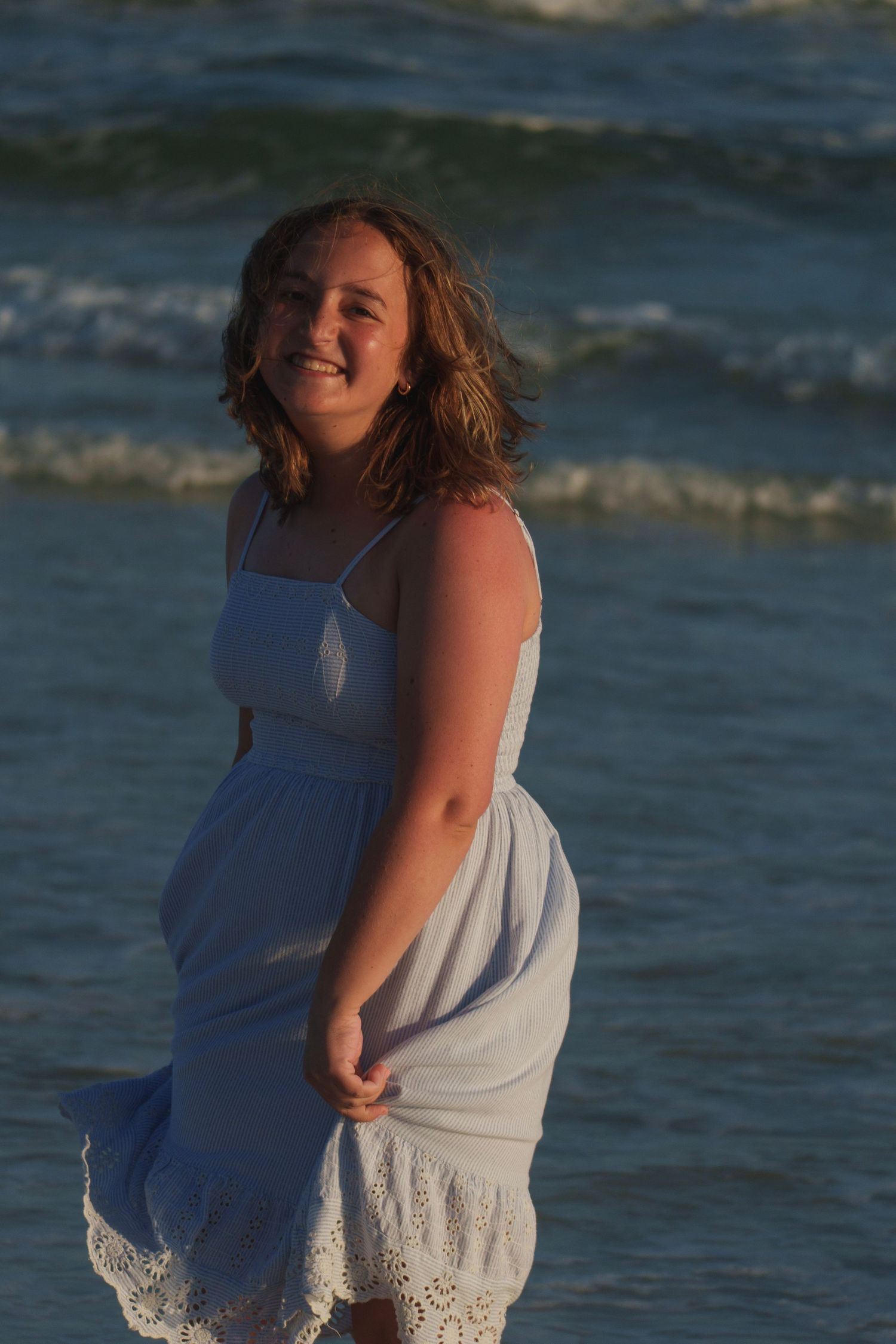 Smiling young woman in a white sundress standing on a sandy beach with ocean waves behind her.