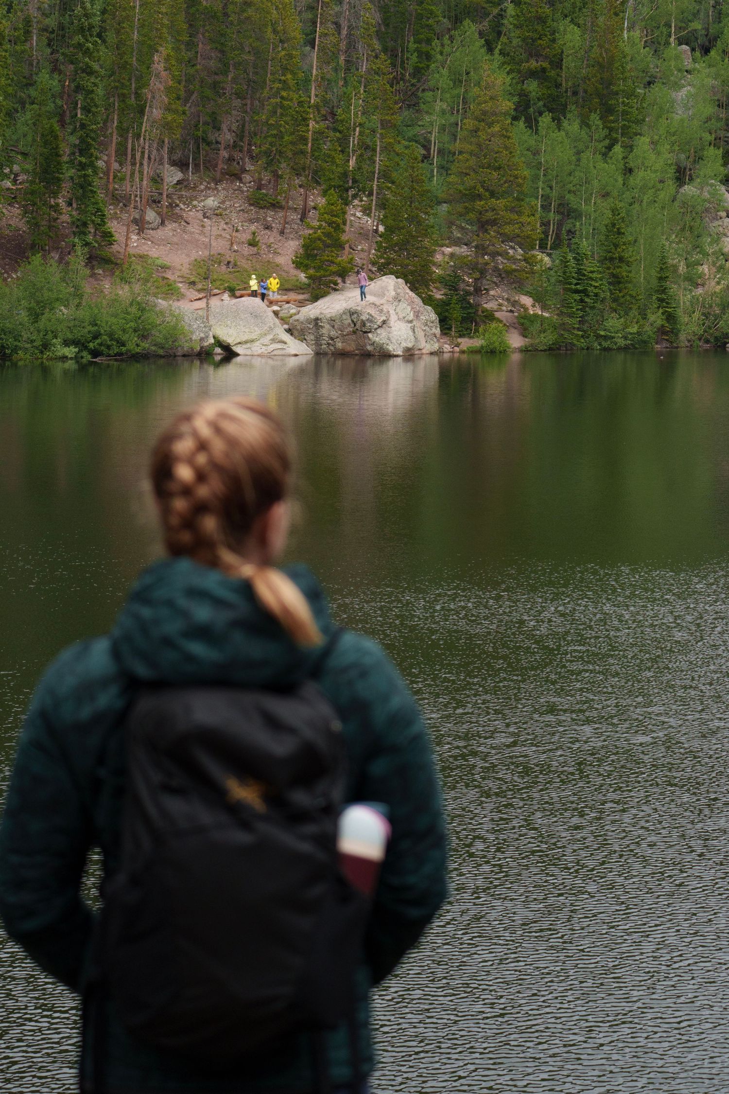 Woman with braided hair and backpack gazing at a calm mountain lake surrounded by pine forest.