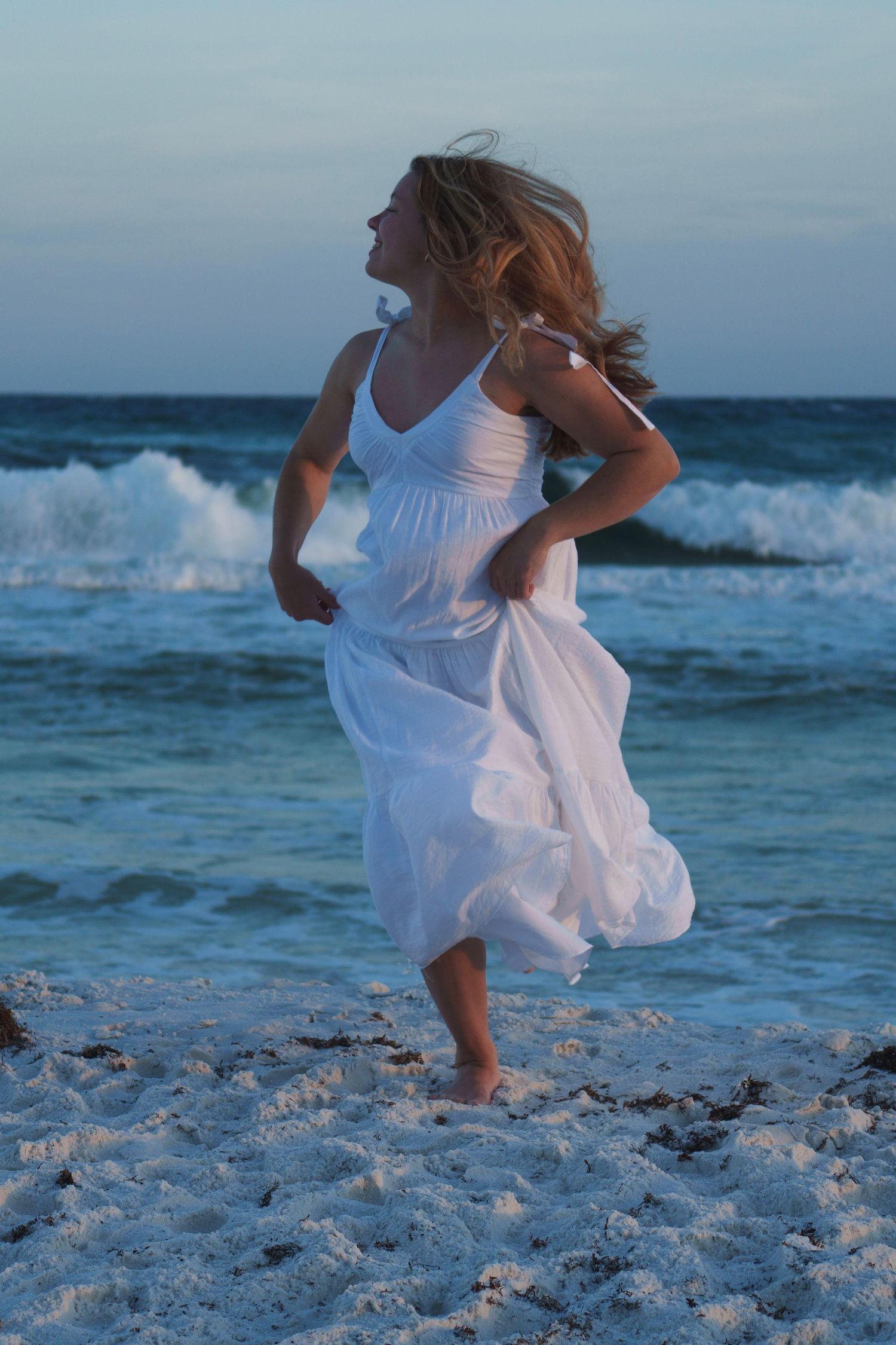 Joyful woman in flowing white dress running barefoot along a sandy beach at dusk.