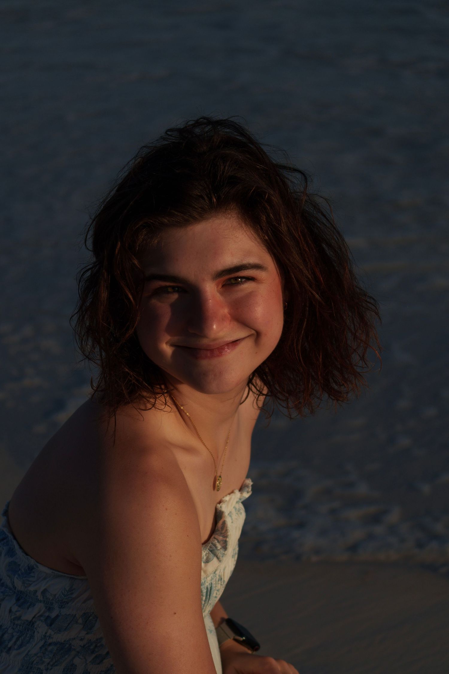 Smiling teenage girl with short curly hair wearing a floral dress sitting on a beach at sunset.