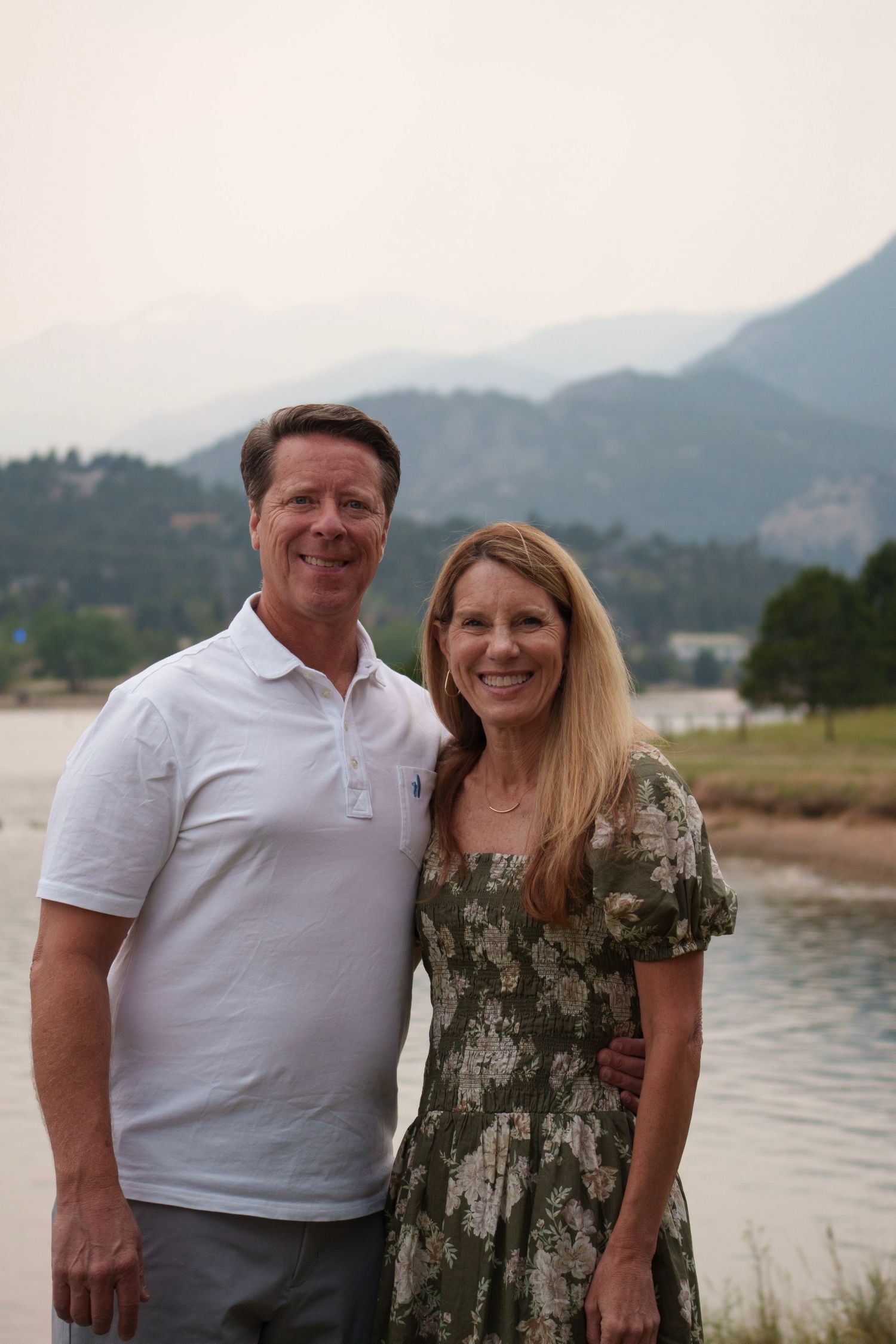Happy couple smiling together by a mountain lake with hazy peaks in the background.