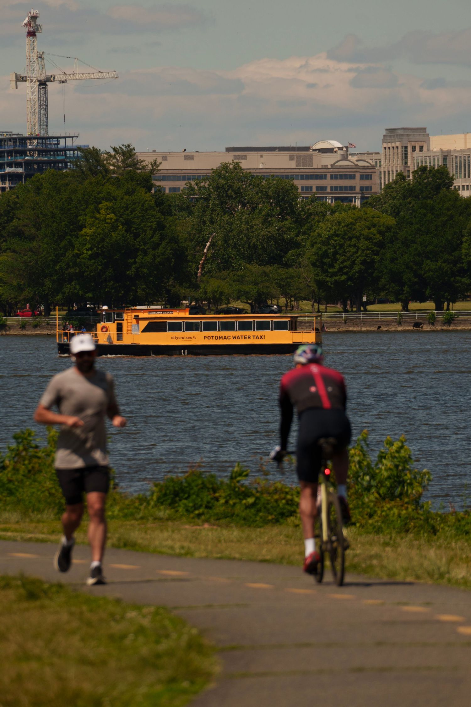 A yellow Potomac Water Taxi ferry crosses the river as a cyclist and jogger pass on a path.